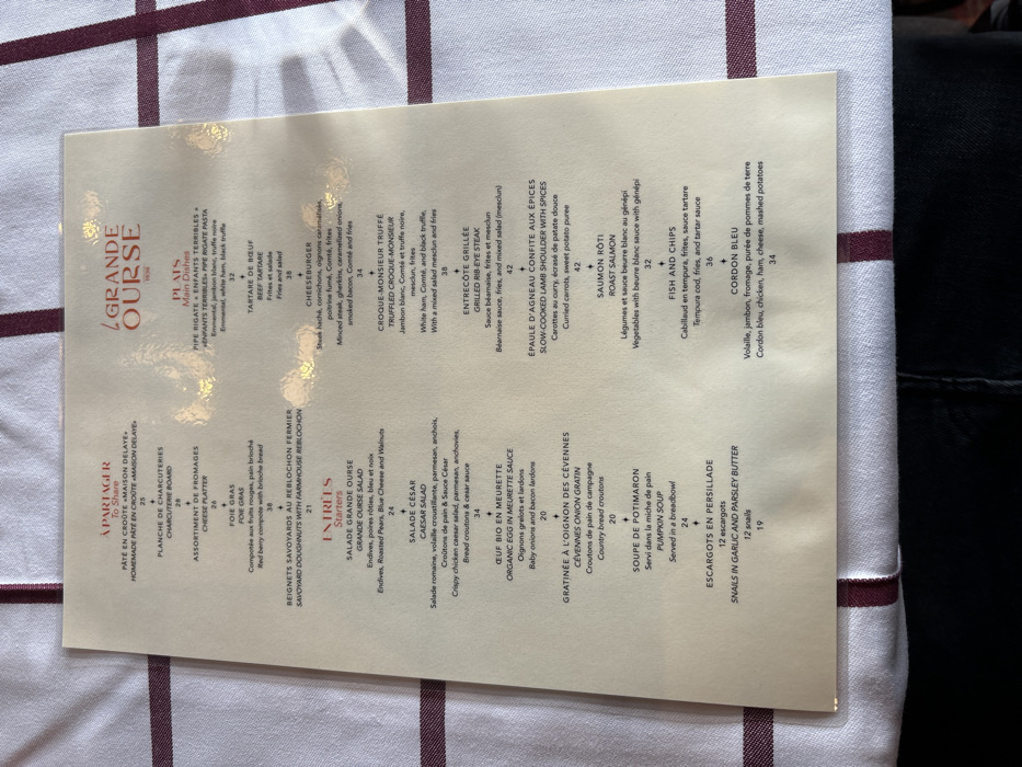 This media file features a restaurant menu, titled "LA GRANDE OURSE 1936," lying diagonally on a white tablecloth adorned with parallel maroon stripes. The setting appears to be indoors, likely a restaurant in Val-d'Isère, France. Glimpses of dark clothing, possibly belonging to a diner, are visible in the top and bottom right corners of the frame.
The menu is presented bilingually in French and English, offering a range of dishes categorized under "À PARTAGER" (To Share), "ENTRÉES" (Starters), and "PLATS" (Main Dishes). Each item includes a brief description and a price in Euros.
Notable items listed include:
* **À PARTAGER:** Homemade Pâté en Croûte (25€), Charcuterie Board (28€), Cheese Platter (26€), Foie Gras (38€), and Savoyard Doughnuts with Farmhouse Reblochon (21€).
* **ENTRÉES:** Grande Ourse Salad (24€), Caesar Salad (34€), Organic Egg in Meurette Sauce (20€), Cévennes Onion Gratin (20€), Pumpkin Soup (24€), and Snails in Garlic and Parsley Butter (19€).
* **PLATS:** Pipe Rigate (32€), Beef Tartare (38€), Cheeseburger (34€), Truffled Croque-Monsieur (38€), Grilled Rib-Eye Steak (42€), Slow-Cooked Lamb Shoulder with Spices (42€), Roast Salmon (32€), Fish and Chips (36€), and Cordon Bleu (34€).
The menu itself has a creamy, light yellow hue with brown text, suggesting a traditional or upscale establishment. Reflections on its surface indicate a glossy finish or protective covering. The clear presentation of dishes and prices suggests a readiness for ordering.