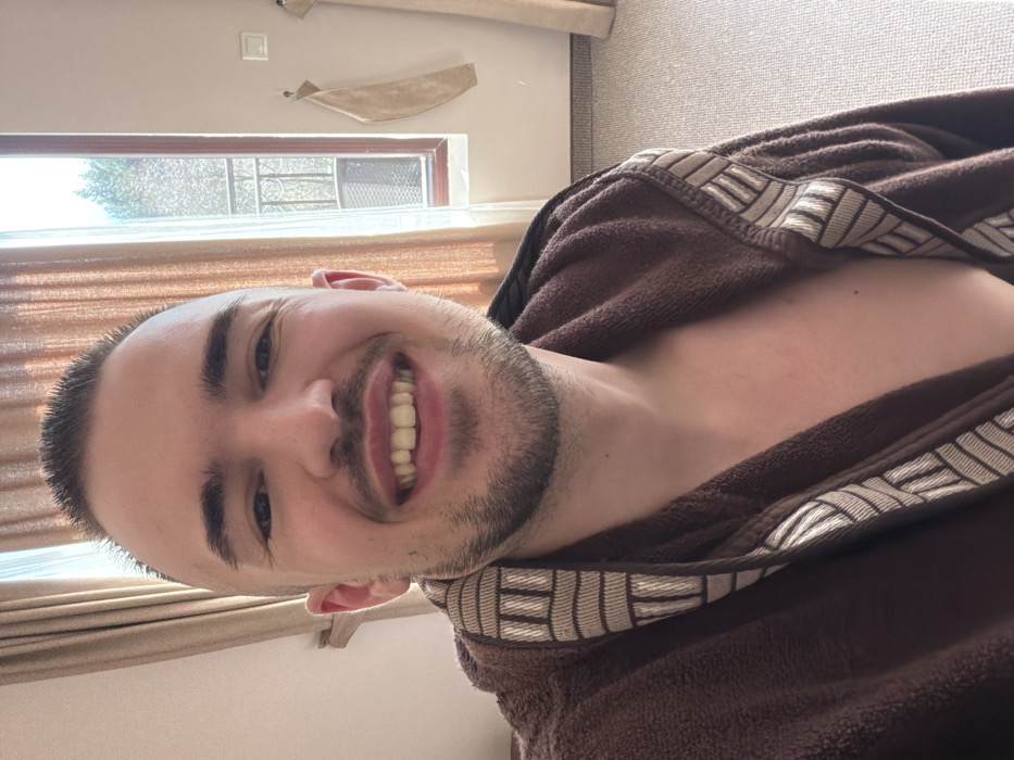 A light-skinned adult male is depicted in a chest-up portrait, smiling broadly towards the viewer. He has a buzz cut hairstyle, dark hair, and light stubble. He is wearing a dark brown, possibly terry cloth, bathrobe with a light-colored geometric pattern on the lapel. His upper chest is exposed. The background is an indoor room in Kladnitsa, Bulgaria, featuring light-colored curtains on both sides. To the right, a window reveals outdoor trees and foliage, with a decorative metal grate visible in the lower section. A light-colored wall section to the right of the window shows two light-colored fabric pieces, possibly towels, hanging on hooks, and a white light switch. The floor is covered with a light-colored carpet. The scene is illuminated by natural light.