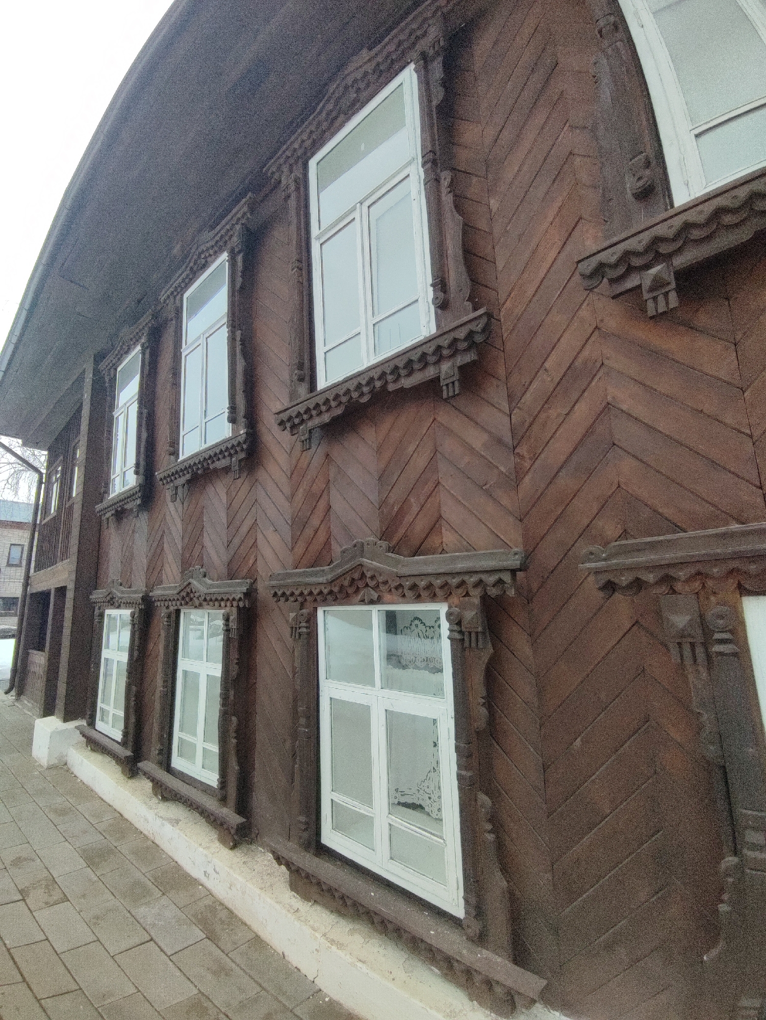 This image shows a detailed view of an old, ornate wooden building, likely located in Sarapul, Russia. The building's exterior is predominantly brown, with a distinctive pattern of chevron-shaped wooden planks on the facade. The building features white-framed windows, which are adorned with decorative wooden carvings and moldings. The windows are arranged in a row, and they appear to have a base of ornately carved wood that sits on a white concrete foundation along the street. The building has a weathered appearance, suggesting age and exposure to the elements. The sidewalk in front of the building is paved with gray stones. In the background, another building is partially visible.