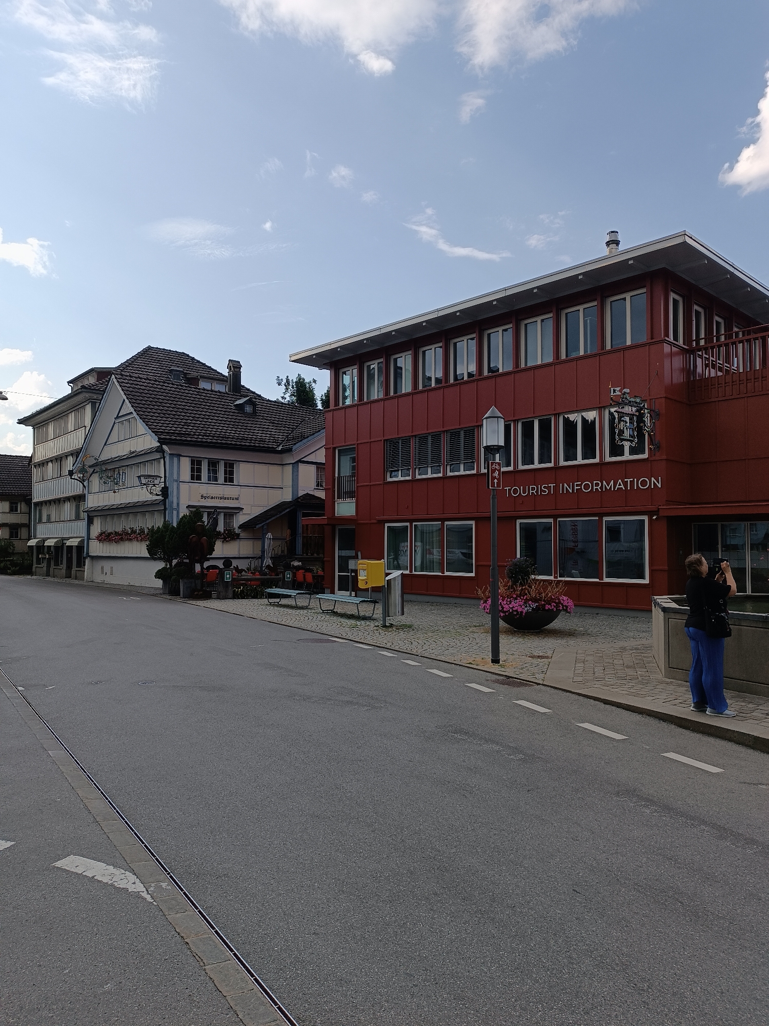 The image depicts a street scene in the city of Appenzell, Switzerland. In the foreground, a paved road runs diagonally from the lower left to the upper right of the frame. A white dashed line marks the edge of the lane closest to the viewer. A drain grate runs parallel to the curb along the right side of the road.
On the right side of the street, a modern, red-colored building with multiple windows stands prominently. Text on the building reads "TOURIST INFORMATION." A woman in blue pants and a dark top is standing near the building, holding a camera and appearing to take a photograph. In front of the building, a planter with pink flowers adds a touch of color, and a stone fountain or water feature is partially visible. A yellow mailbox is also present near the building's entrance.
Adjacent to the modern building and further to the left is a more traditional, multi-story building with a pitched roof. This building has a cream-colored facade with white trim and dark brown tiled roofing. Signage on this building indicates it is a "Speiserestaurant" (restaurant). Benches are placed outside this establishment, suggesting an outdoor seating area.
Further down the street to the left, other buildings with similar traditional architectural styles are visible. The sky above is blue with scattered white clouds, indicating fair weather. The overall scene suggests a pleasant day in a town with a mix of modern and traditional architecture.