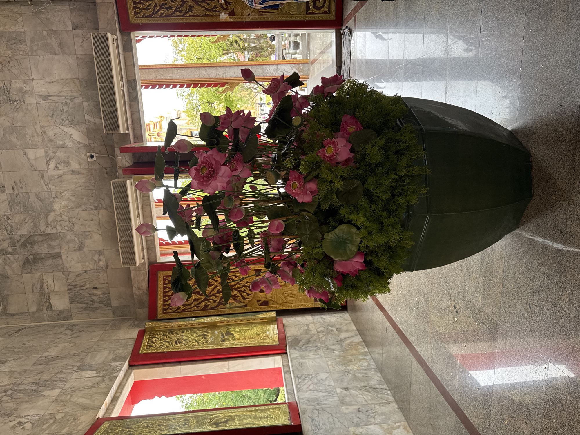 A large, green, polygonal planter filled with artificial pink lotuses and lush green foliage sits on a polished granite floor inside a building in Chalong, Thailand. The planter is positioned in an entryway with ornate gold and red doors opening to an outdoor area with trees and a structure resembling a temple. The walls of the interior are clad in grey marble tiles, and two air conditioning units are mounted high on the wall. Sunlight streams in from the open doorways, creating reflections on the floor. The overall scene suggests a temple or ornate public building in a tropical setting.