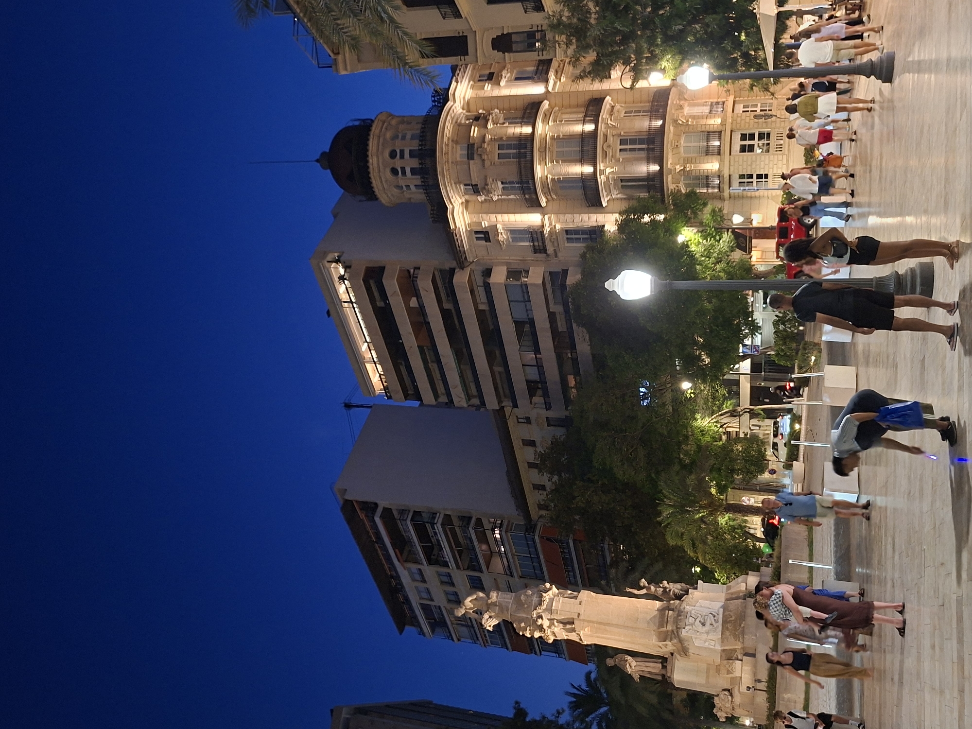 The image depicts a night scene in the city of Alicante, Spain, specifically at the Explanada de España. In the foreground, several people are visible, including a woman holding a child, a man bending down to pick up a glowing object, and a couple standing near a lamppost. The background features a monument on the left and elegantly lit buildings on the right, including a distinctive building with a domed tower. Palm trees and streetlights add to the ambiance. The overall setting suggests a public square or promenade, bustling with activity in the evening.