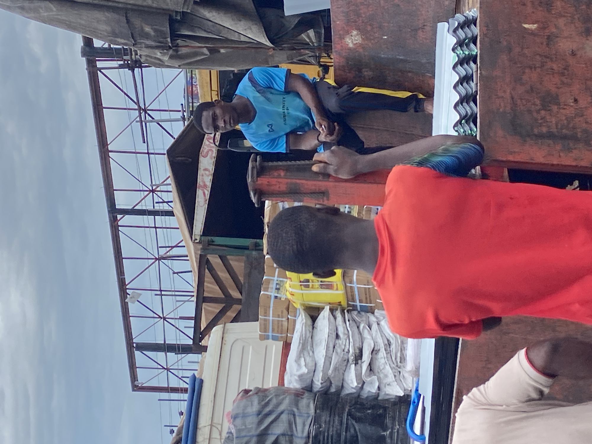 The image depicts three individuals in an outdoor setting, likely in Ogidi, Nigeria. The scene appears to be a loading area, with a truck visible in the foreground laden with goods. Two men are the primary subjects. One man, wearing a blue t-shirt with a logo and black pants, is seated on a red metallic object, possibly part of the truck's loading mechanism. He is looking towards the viewer with a neutral expression. Another man, whose back is mostly towards the camera, is wearing a bright orange t-shirt and appears to be interacting with the seated man. A third person, wearing a light-colored shirt with red trim, is partially visible in the lower right corner.
The truck is loaded with various items, including boxes wrapped in brown paper and secured with blue straps, and several white sacks. The corrugated metal roofing of a nearby structure is visible, suggesting a covered market or storage area. The overall atmosphere seems to be one of commerce or logistics. The lighting suggests it is daytime, with overcast skies. There is no discernible text in the image, apart from a logo on the blue t-shirt which is not clearly legible. The context of Ogidi, Nigeria, places this scene in a West African urban or peri-urban environment.