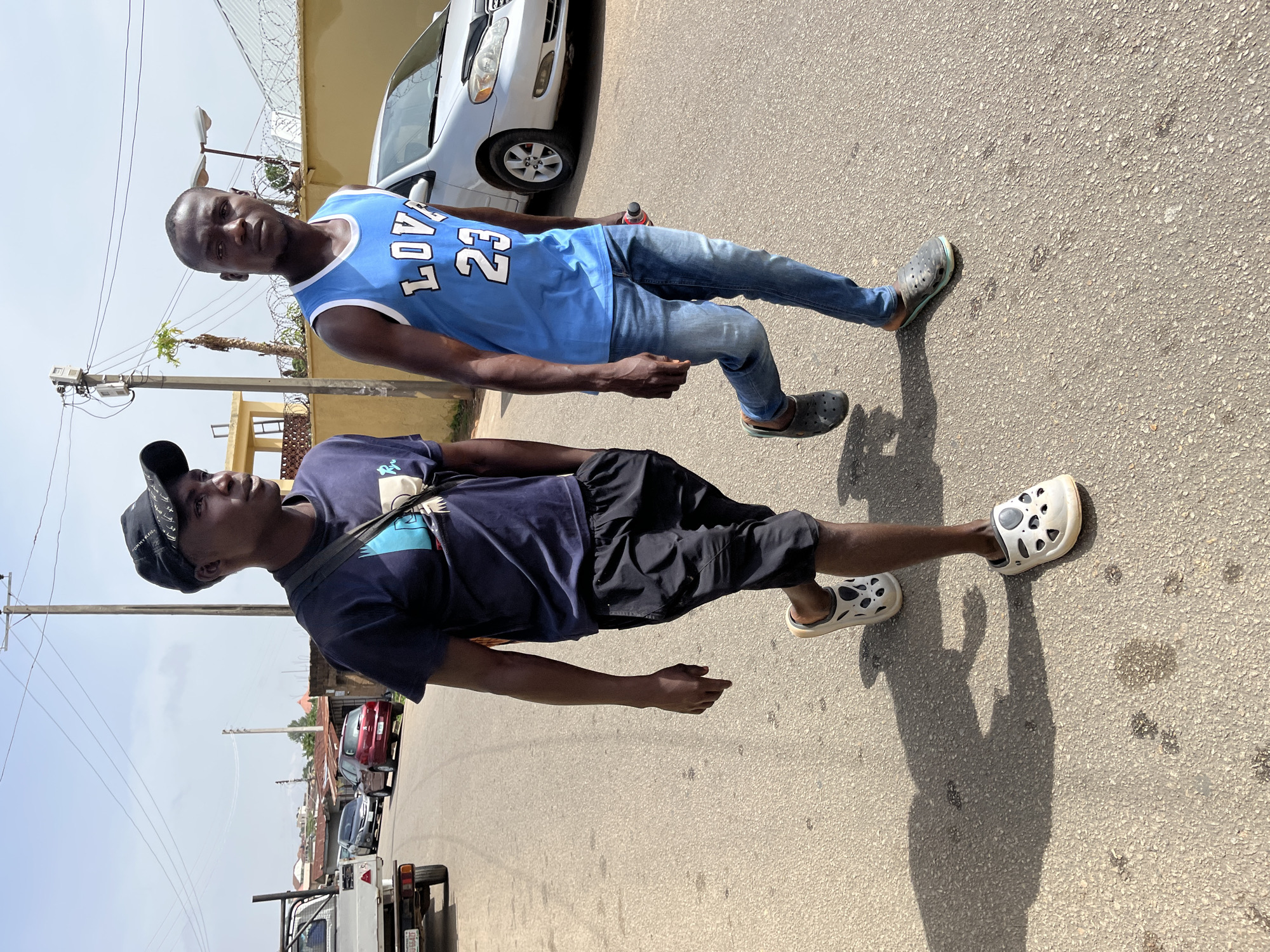 Two young African men are standing on an asphalt road. The man closer to the foreground wears a navy blue t-shirt, black shorts, and white Crocs. A black baseball cap is on his head, and a strap crosses his chest. The other man wears a blue jersey with white trim, blue jeans, and dark-colored Crocs. A silver car is partially visible in the background. Utility poles and wires line the street. Other vehicles, including a white pickup truck and a red car, are parked further down the street. Buildings with corrugated iron roofs are visible in the distance. The photo appears to have been taken during the day in Jos, Nigeria.