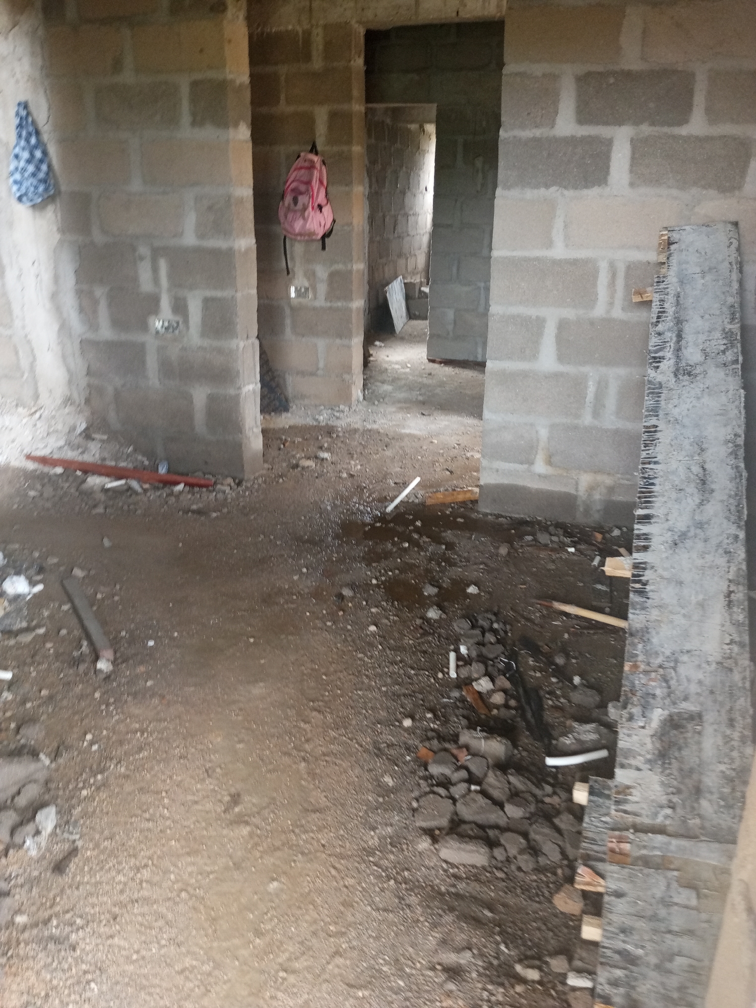 The image shows the interior of a building under construction in Jalingo, Nigeria. The walls are made of concrete blocks, and the floor is a mixture of dirt and gravel, with some areas appearing wet or muddy. There are several doorways leading to other parts of the building, which are also unfinished.
Objects visible include a blue and white patterned cloth hanging on the left wall, and a pink backpack hanging on a wall in the center of the image. There are also some construction materials scattered on the floor, such as a red pipe, pieces of wood, and rubble. A long, grey plank of wood is leaning against a wall on the right side of the frame.
The lighting suggests it is daytime, but the weather is unclear. The scene depicts a raw construction site, implying that work is either ongoing or has recently paused. There is no visible text in the image. The overall impression is one of an incomplete structure in a tropical or sub-tropical environment, given the location context.