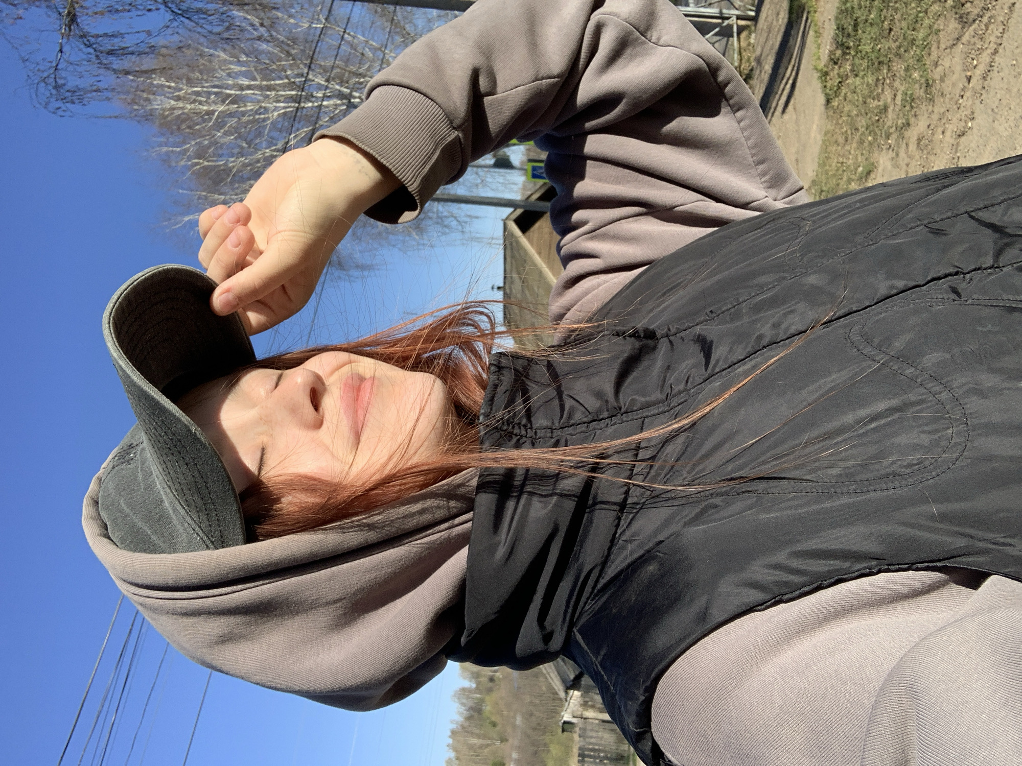 A medium shot presents a person outdoors in what appears to be Фоки, Russia. The individual has fair skin and red hair, wears a gray hoodie under a black vest, and is holding the brim of a gray cap. The person's eyes are closed. The background shows a clear blue sky, bare trees, and a glimpse of nearby buildings and greenery. A road is barely visible in the far background.