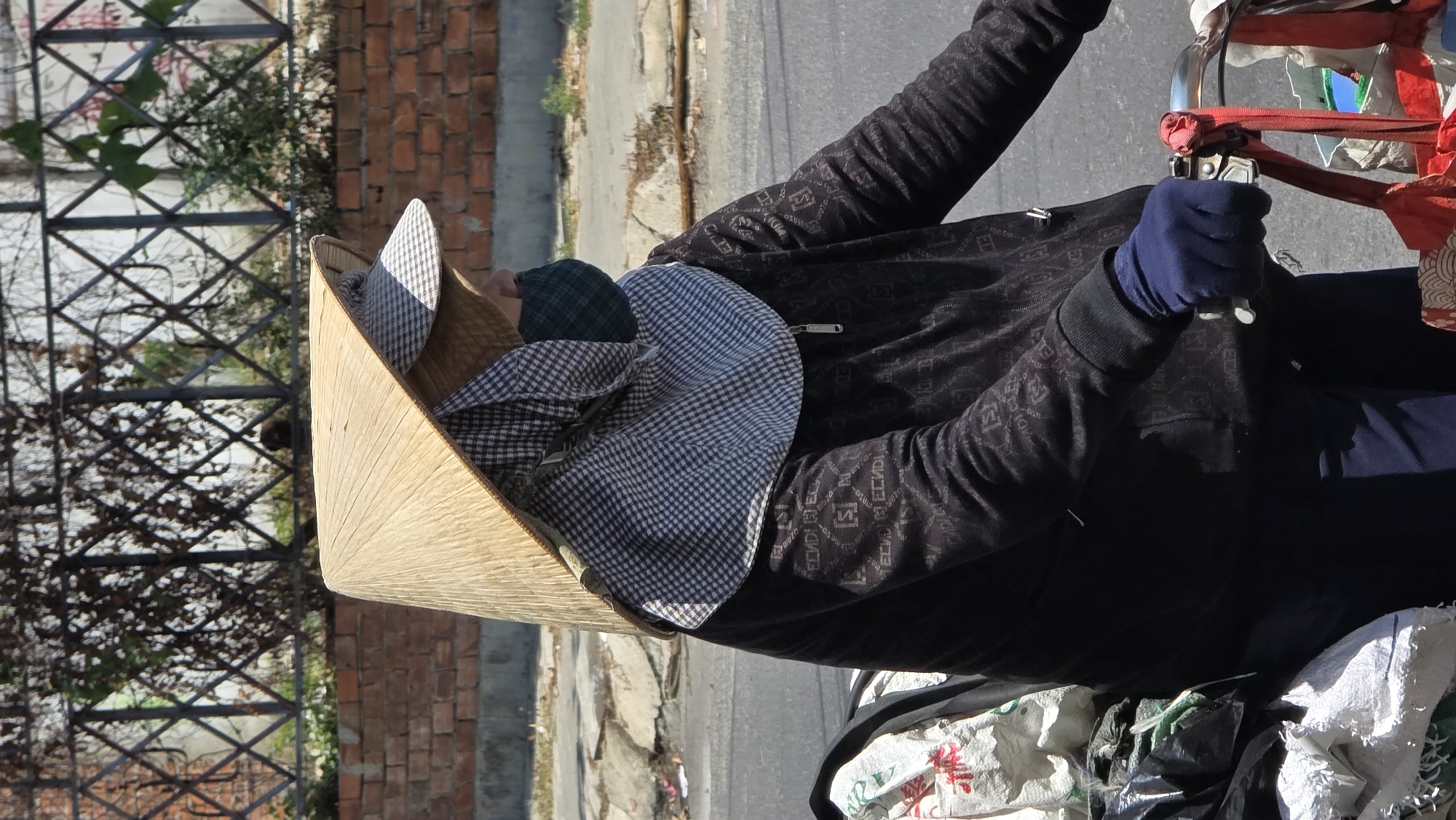 A person wearing a conical Asian hat and dark clothing operates a vehicle on a city street, possibly in Bắc Nha Trang, Vietnam. The person wears blue gloves. Bags of items are on the front of the vehicle. The background shows a sidewalk, a brick wall, and a metal fence.