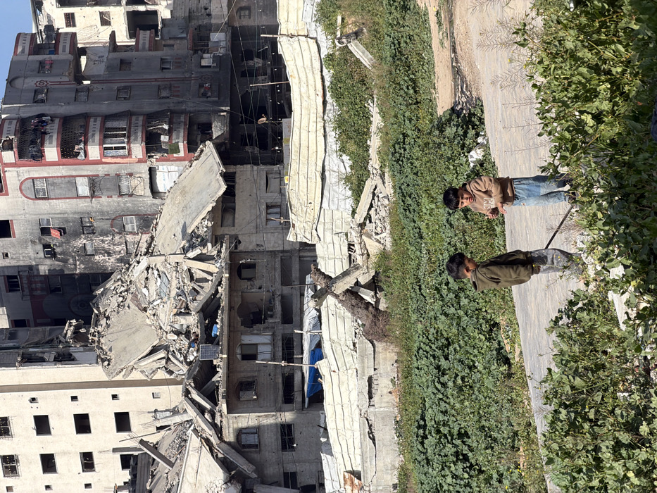 A severely damaged high-rise building stands amidst rubble. Sections are collapsed, and windows are destroyed, revealing a dark, charred interior. Two boys stand on a sidewalk next to dense green bushes. One wears a brown jacket and jeans, while the other is in a green jacket. Debris is visible on the ground near the bushes, and the building's ruins cast a long shadow over the scene. The overall impression is one of devastation and displacement.