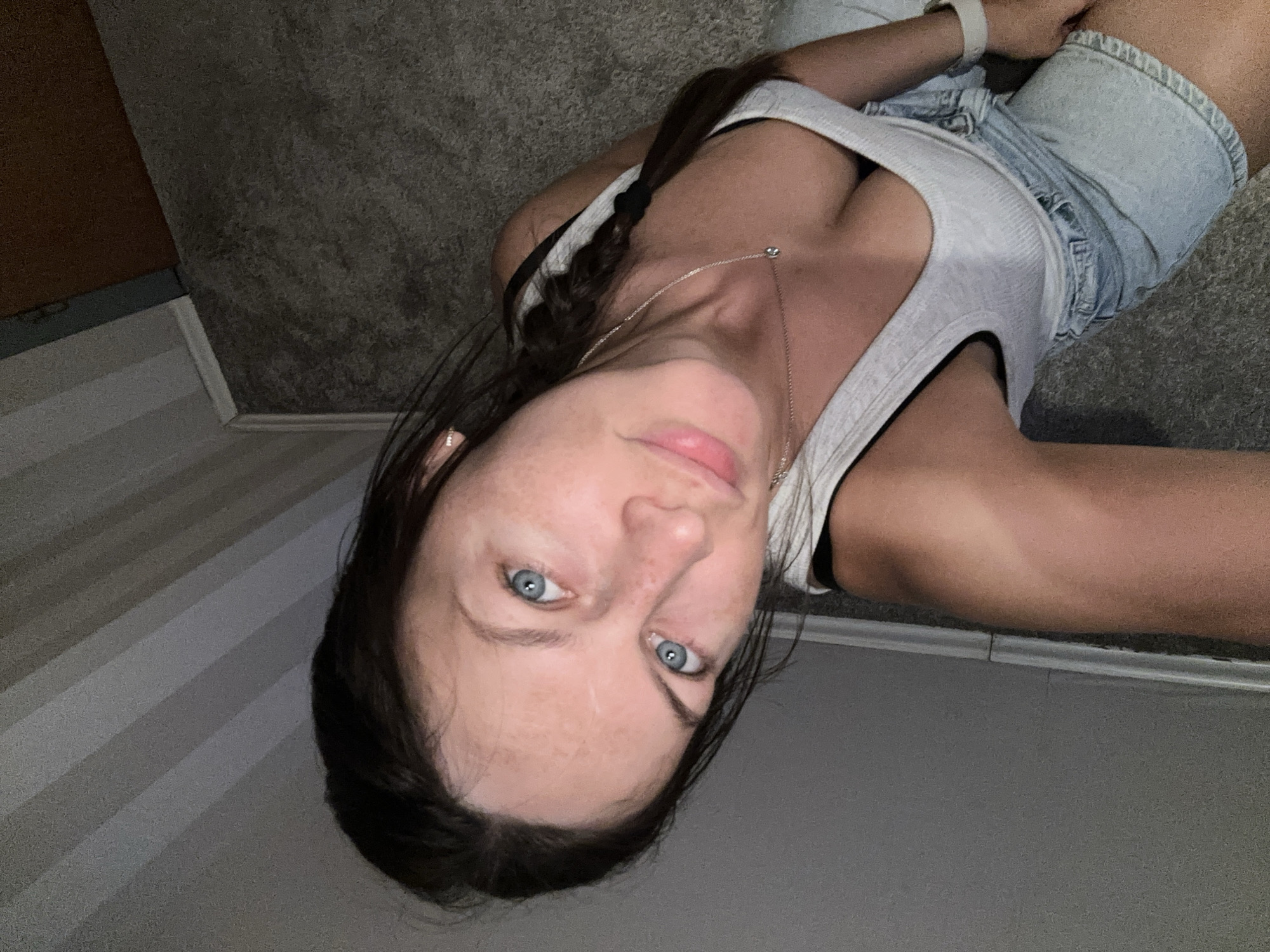 A young woman is depicted in a close-up, upward-angled shot, lying down indoors. She has long, dark hair styled in a braid that rests over her shoulder. Her striking blue eyes gaze directly towards the camera, and her face shows a light complexion with visible freckles on her forehead and nose. Her lips are slightly parted.
She is wearing a light-colored (possibly white or light grey) ribbed tank top or camisole, revealing a glimpse of a dark bra strap. A delicate silver chain necklace with a small pendant adorns her neck. Below her torso, she wears light blue denim shorts. A white wristband, likely a watch or fitness tracker, is visible on her left wrist.
The setting appears to be a room, with the woman lying on a dark grey, textured surface, likely carpet or a rug. Parts of a baseboard and a wall, possibly a door frame, are visible in the background, suggesting an indoor environment. The lighting is somewhat dim, implying evening or low-light conditions. The image itself is rotated, giving the impression of an unconventional perspective. No visible text is present. The overall scene is casual and personal, located in Fredenbeck, Germany.
