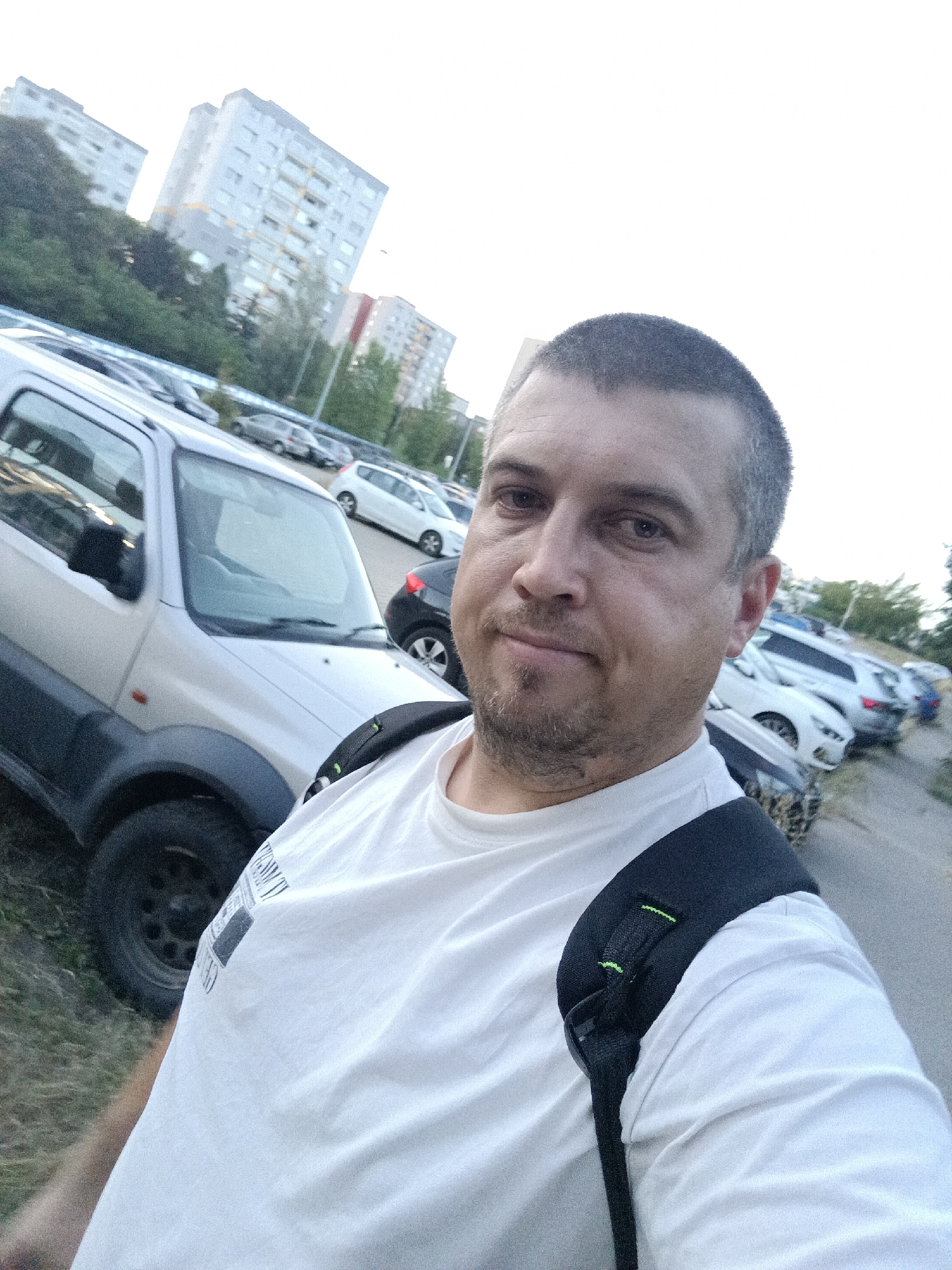 A man takes a selfie in a parking lot with a silver Suzuki Jimny in the foreground. The man is wearing a white t-shirt and a black backpack. In the background, several cars are parked, and apartment buildings and trees are visible. The sky is overcast. The image was taken at dusk. The location is the parking lot of the shopping center Galaxie on the border of Prague 5 and Prague 13, Czech Republic.