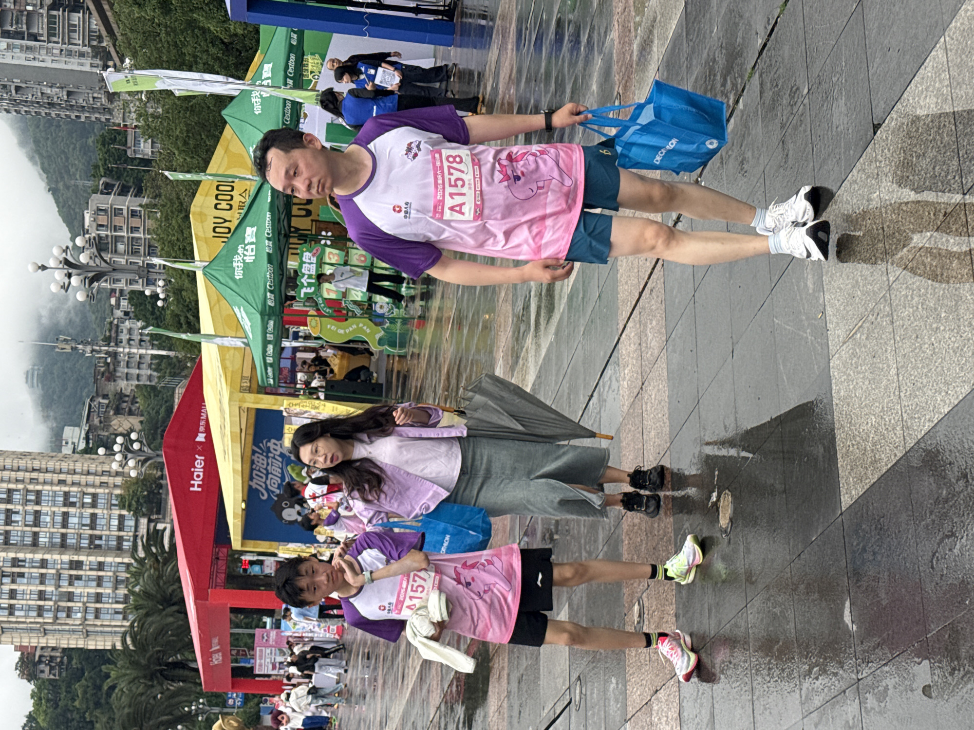 Here's a detailed description of the image:
**General Overview:** The image depicts a scene on a wet, paved area, likely a plaza or walkway, with buildings and greenery in the background. It appears to be a snapshot taken during or after a running event.
**People:** There are several people visible:
* A man wearing a white and purple running shirt with the number "A1578", blue running shorts, and white sneakers is carrying a blue Decathlon bag.
* A woman is holding an umbrella and wearing a light purple jacket and a gray skirt.
* Another person wearing a running shirt similar to the man and pink sneakers.
**Objects:**
* Blue Decathlon bag
* An umbrella
* Several tents and banners with branding (Haier, Joy Cool, Cestbon)
* Race number tag "A1578"
* Street lamps
**Activities:**
* People are standing around, possibly recovering or waiting after a running event.
* The woman is holding an umbrella, indicating it has been or is raining.
**Environment:**
* The location seems to be in a city. Buildings are visible in the background.
* The ground is wet, suggesting rain has recently fallen.
* There's greenery in the background, including trees.
**Location (Based on OCR):**
The race number on the shirt includes the characters 2026 重庆六一, indicating that the image was taken in the city of Chongqing, likely during a race with a date related to "June 1st". Given "Nan'an District" is indicated in the prompt, the location would likely be within that district of Chongqing, China.