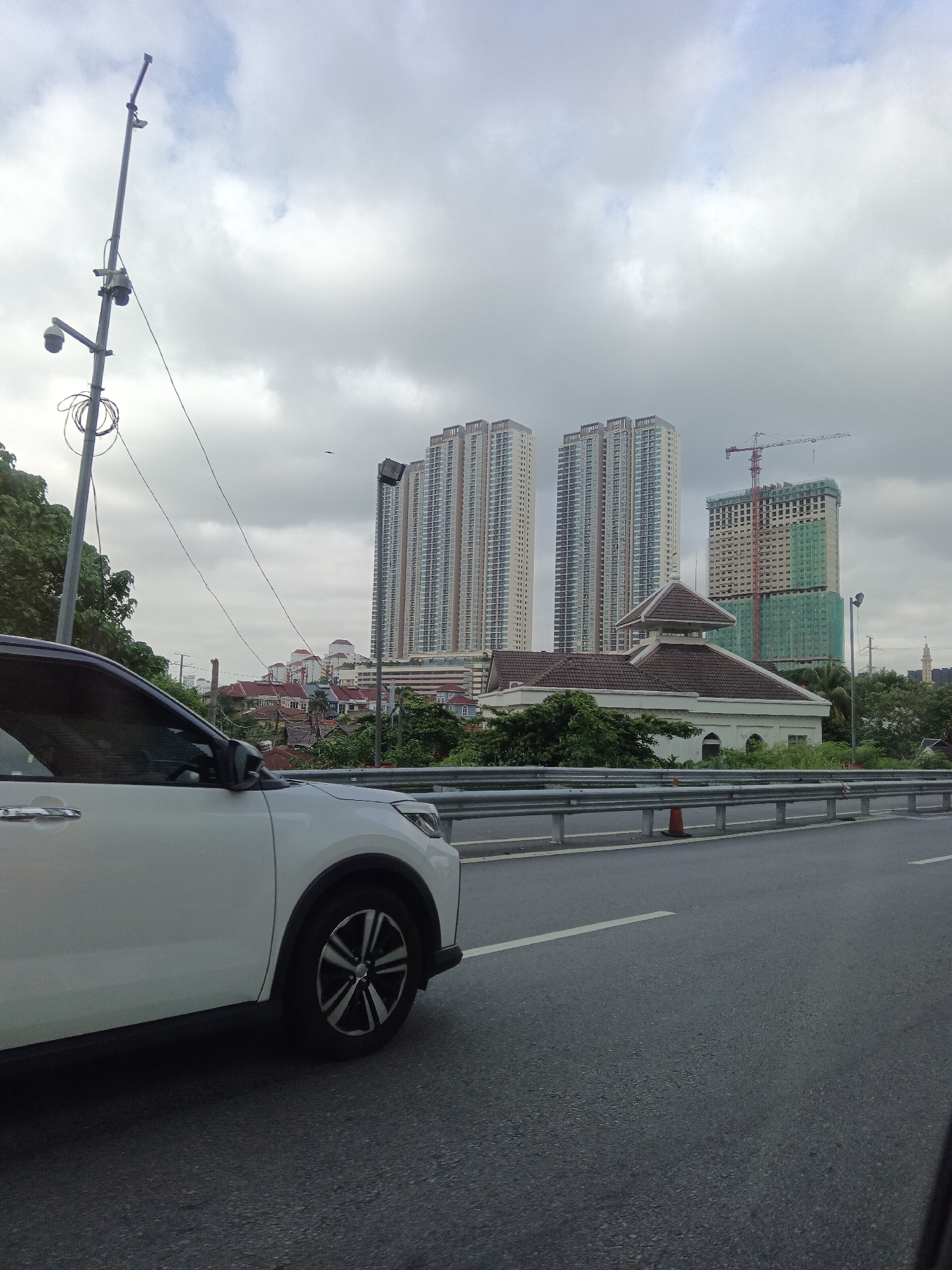 The image depicts a view from a vehicle driving on a highway in Kuala Lumpur, Malaysia. In the foreground, the front passenger side of a white car is visible. The highway features a grey asphalt road surface with a white lane marking. A metal guardrail runs along the side of the road, with a traffic cone visible behind it.
In the background, a cluster of buildings forms the urban landscape. Three tall, modern residential towers with a light-colored facade and numerous windows dominate the skyline. To the right of these towers, a building under construction is covered in green netting, with a construction crane extending upwards.
In front of the residential towers, a lower-rise building with a brown tiled roof and a domed central structure is visible. This building appears to be a mosque or a similar religious or cultural establishment. A street lamp with multiple cameras is situated to the left of the frame. Dense green foliage lines the sides of the highway and surrounds the buildings. The sky is overcast with grey clouds. The scene suggests an active urban development environment with both completed and ongoing construction projects.