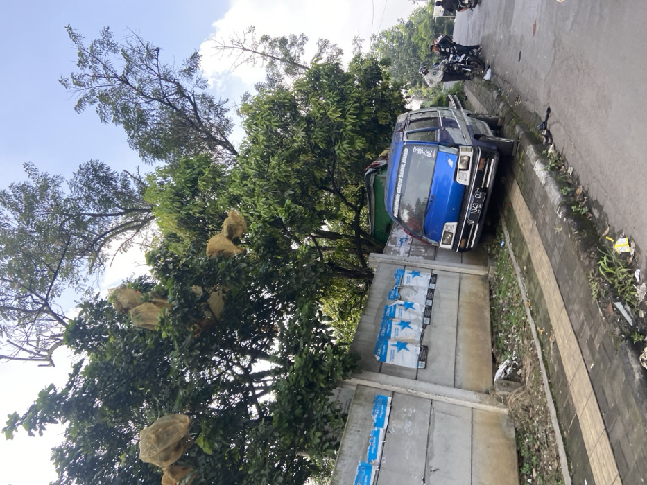 The image shows a blue minivan parked on the side of a paved road in Kecamatan Cimahi Selatan, Indonesia. The vehicle is parked facing forward with its front end closest to a concrete wall with a sidewalk between the wall and the road. There are several posters attached to the wall. The van is in a residential area and a large tree is in the foreground to the left of the vehicle, with a smaller tree to the right. The van is parked partially on the sidewalk and partially on the road. There are three people in the distance, all dressed in dark clothing, who are walking away from the van. The ground is covered in loose debris. The sky is clear and the sun is shining. The coordinates provided are -6.93 latitude and 107.565 longitude.