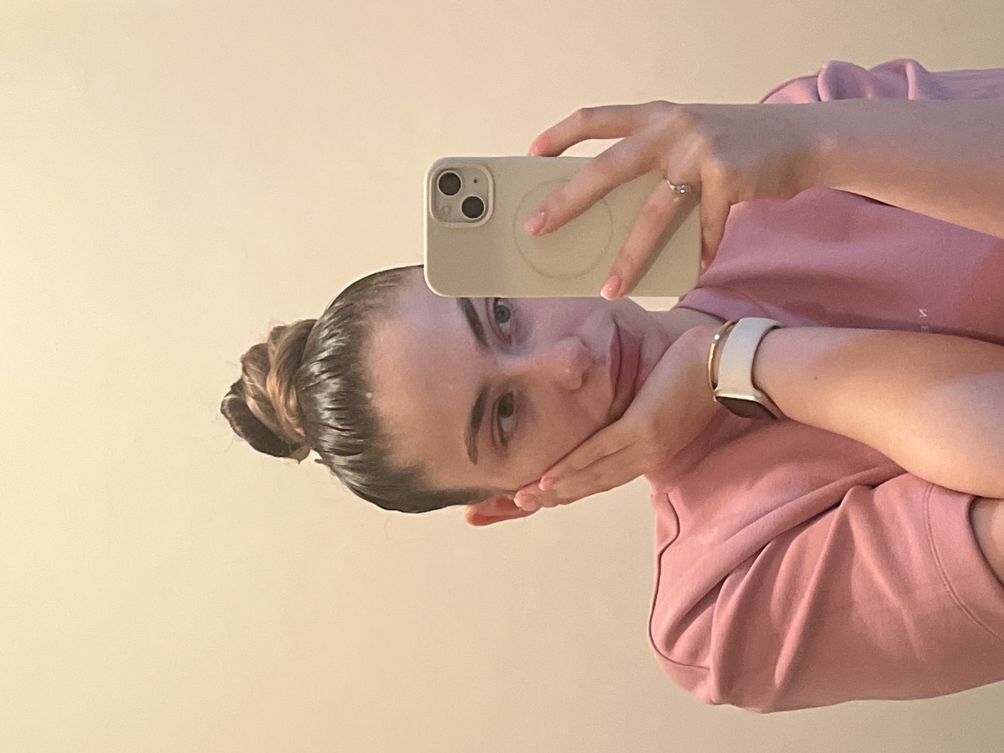 A young woman is captured in a mirror selfie, with the image rotated 90 degrees clockwise. She has her hair pulled back into a neat, slicked-back bun, giving it a slightly wet or polished appearance. Her face, seen from a slight upward angle, is turned towards the camera, and her left hand is gently resting against her cheek.
She is wearing a soft pink long-sleeved top or sweatshirt. On her left wrist, she wears a smartwatch with a white band and a metallic, possibly gold-toned, casing. Her right hand, holding a light-colored smartphone, is positioned in front of her face. The phone appears to be an iPhone model, featuring two distinct camera lenses arranged vertically and a circular MagSafe ring visible on the back of its case. A simple ring with a small, clear stone is worn on the ring finger of her right hand, and her fingernails have a light pink polish. The background is a plain, light-colored wall, indicating an indoor setting. There is faint, illegible text visible on her pink top.