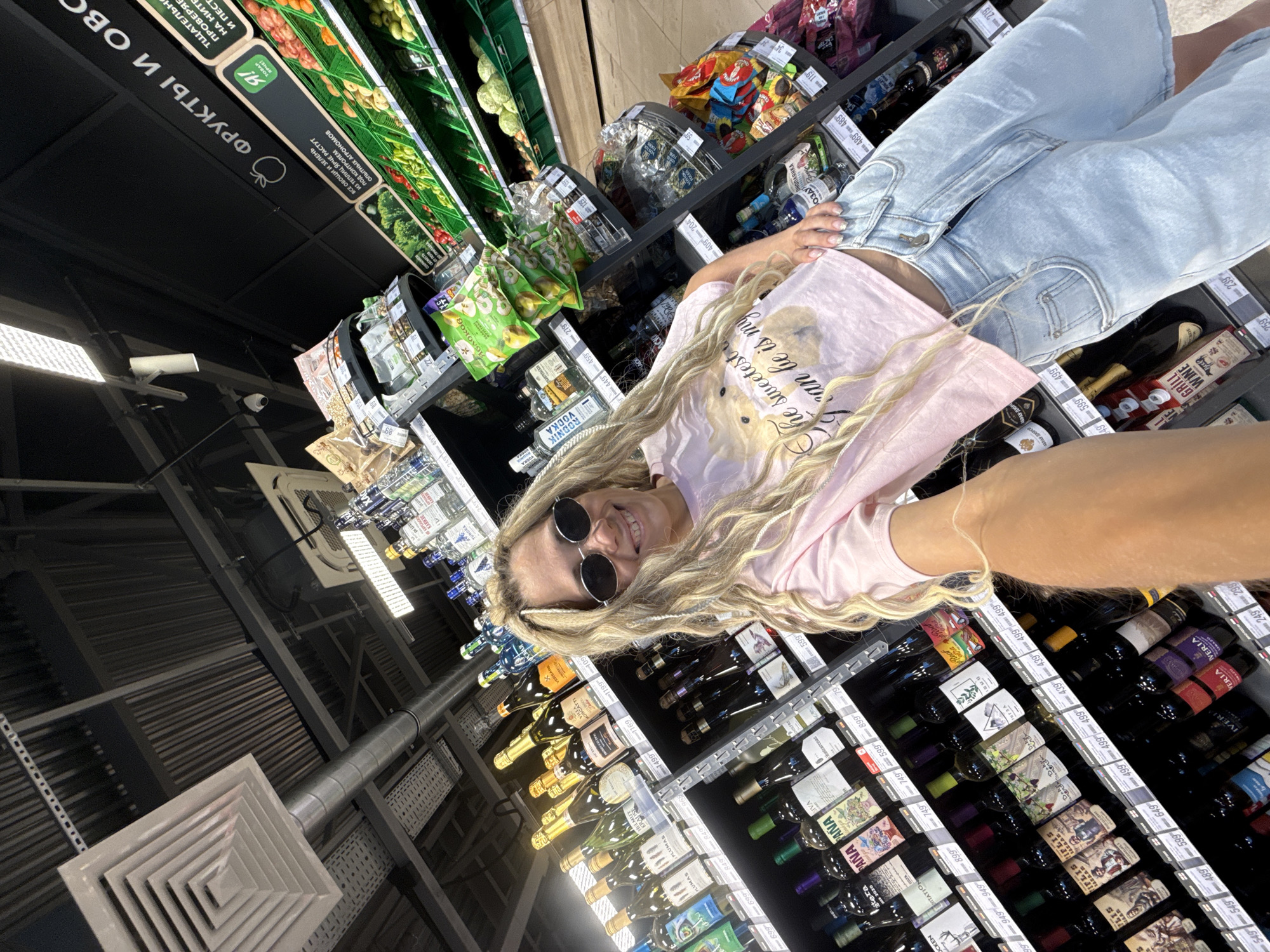 The image shows a woman with long, blonde hair wearing sunglasses in a supermarket setting. She's dressed in a pink top and light blue jeans, and she appears to be taking a selfie in a store aisle, focusing on shelves stocked with wine bottles. The foreground is dominated by various bottles of wine with price tags. In the background, shelves with snacks and vegetables are visible, along with supermarket signage in a language other than English. The location seems to be in a supermarket, potentially in the city of Rusanovo, Russia, based on possible references in the signage text. The photo has been taken at an angle.