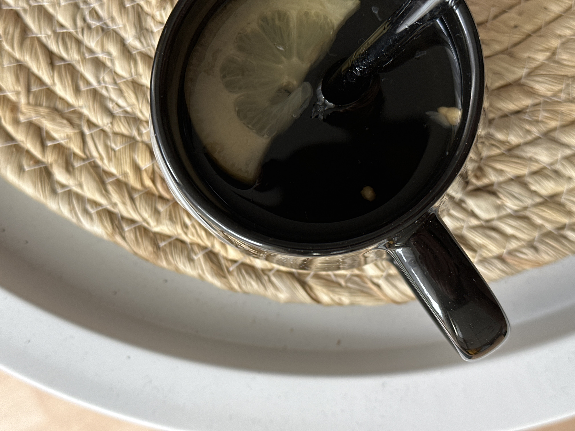A close-up overhead shot shows a mug of dark liquid with a slice of lemon and a metal straw, resting on a woven placemat. The placemat is circular and made of light brown, woven material. To the left of the placemat is the edge of a white tray. The mug is black and appears to be made of ceramic, with a shiny finish. The liquid inside is dark and opaque, with a visible slice of lemon floating on top. The lemon slice is pale yellow and shows its distinct segments. The metal straw is dark and appears to be made of stainless steel. Tiny specks of what might be tea leaves or other small particles are visible in the liquid. The lighting suggests natural light coming from the side, creating subtle shadows and highlights on the mug and straw. The background is blurred, with a hint of a light-colored surface that could be a table. The scene is static and appears to be indoors, likely in a domestic setting. The location is Unknown, Unknown.