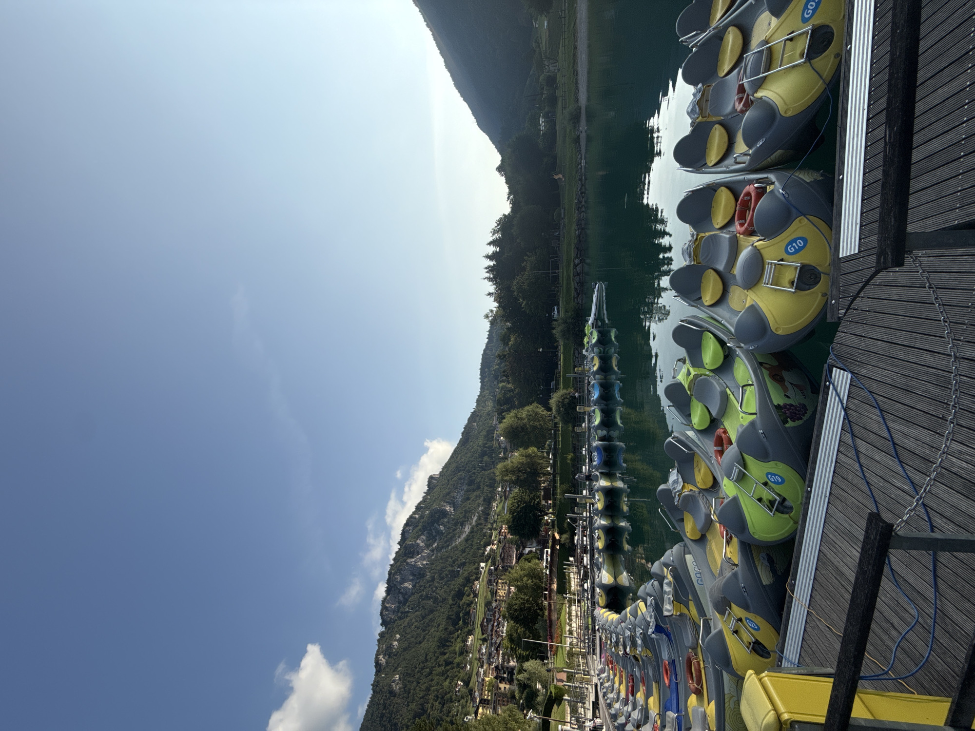 The image shows a row of small boats docked on a wooden pier, with a calm lake and a village nestled at the base of a mountain range in the background. The boats are primarily yellow and grey with green seating, and some have life preservers attached. The sky is a clear blue with a few wispy clouds. The village appears to be a tourist destination, with buildings of various sizes and colors visible on the hillside. The overall scene suggests a peaceful and scenic location, likely in a mountainous region of Italy, possibly Molveno given the context. There are no people visible in the immediate foreground or on the boats, but a person can be seen standing on the pier in the distance. The water of the lake is a deep, clear turquoise, reflecting the surrounding landscape and the sky.