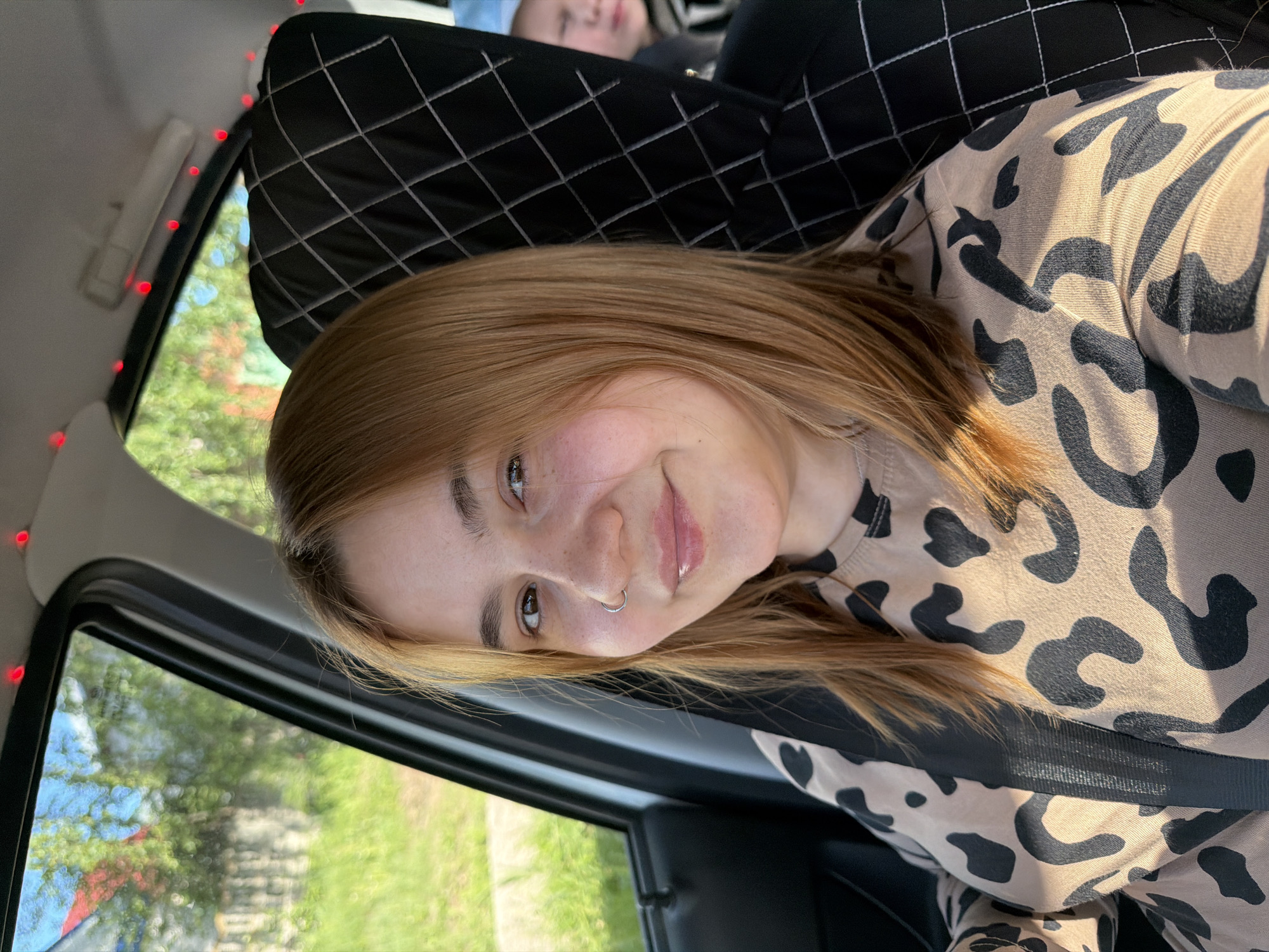 A woman with strawberry blonde hair and a nose ring is in the passenger seat of a car. She is wearing a black and tan animal print shirt and a seatbelt. The car has red lights along the interior roof near the window. The seat has a quilted black seat cover. A second person is visible in the background. Outside the car is greenery and possibly a building. The location may be Chaikovsky, Russia, but this is not verifiable from the image.