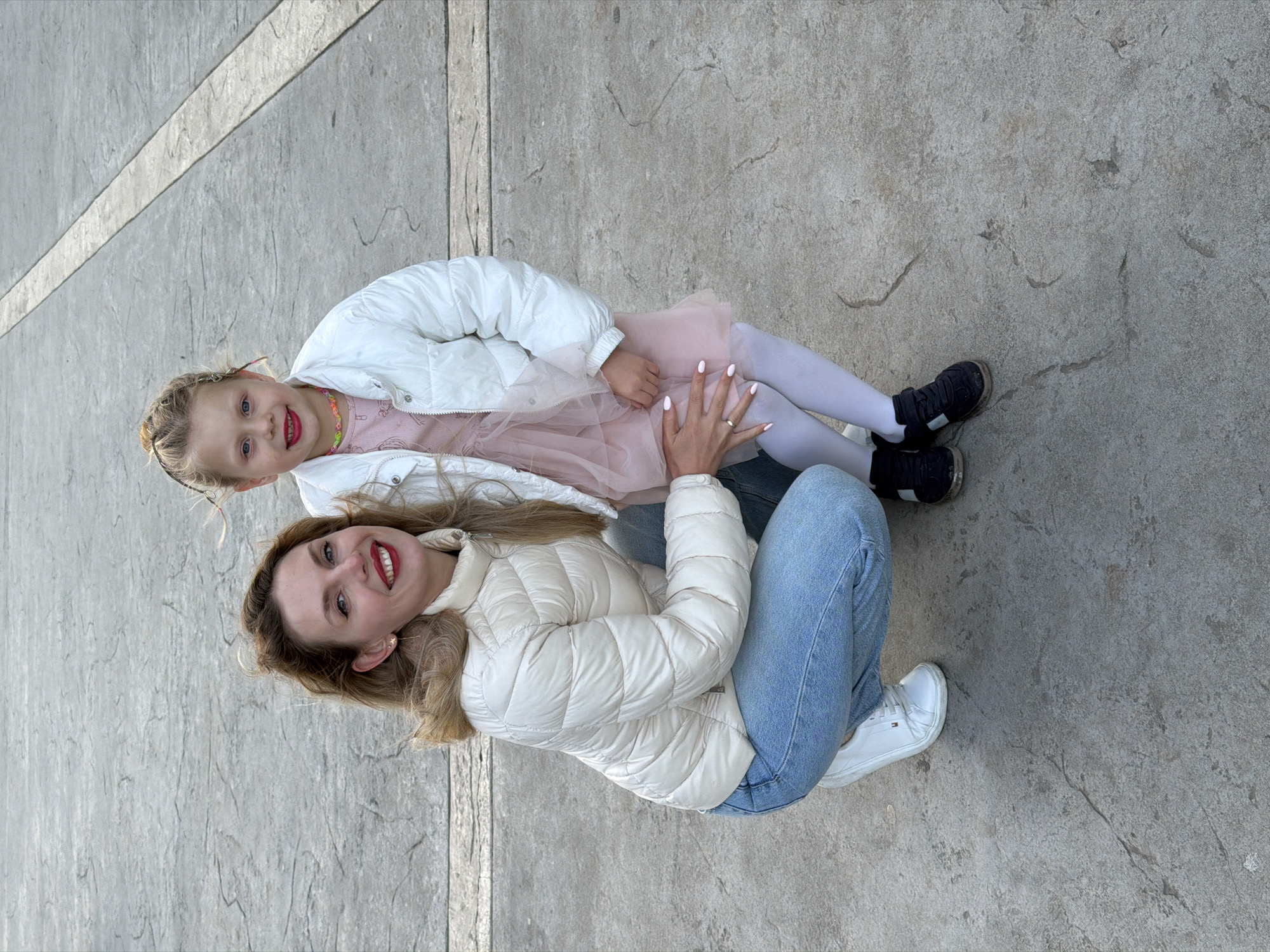 A young girl and a woman are sitting close together on a concrete surface in Budva, Montenegro. The girl is wearing a pink dress, white tights, a white jacket, and dark shoes. The woman is wearing light blue jeans, a white jacket, and white sneakers. Both are smiling at the camera. The concrete surface has a geometric pattern of light-colored lines. The photo is taken from a high angle.