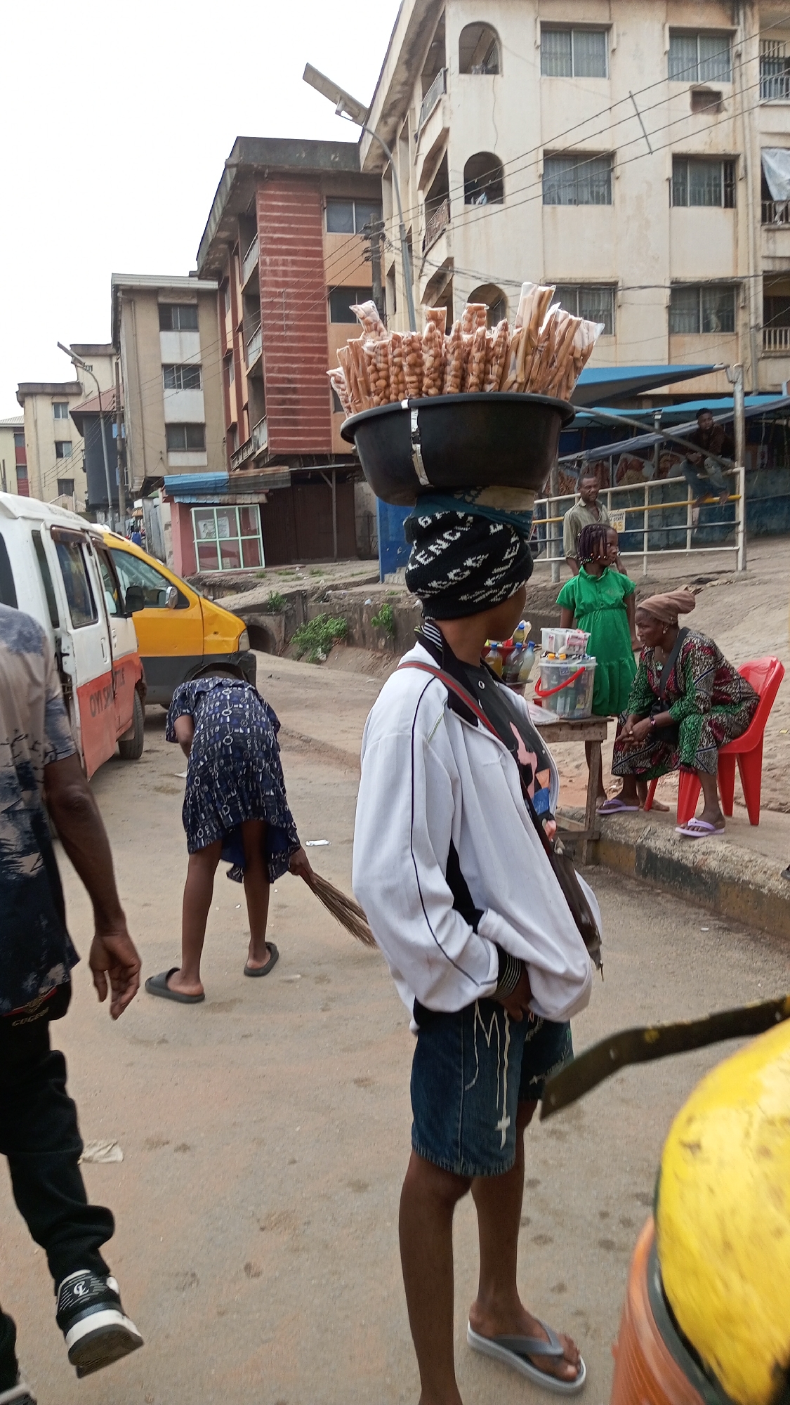 The image captures a street scene in Onitsha, Nigeria. A woman balances a large bowl filled with bagged items, possibly snacks, on her head. She wears a patterned head covering, a white jacket, denim shorts, and sandals. She has a shoulder bag. In the background, a girl in a green dress stands behind a makeshift table where an older woman wearing a headwrap and a colorful outfit sits on a red plastic chair. Another girl is sweeping the street. A white van with a yellow stripe is partially visible on the left. Several buildings are in the background. The street surface is dusty and shows signs of wear. The weather appears overcast.