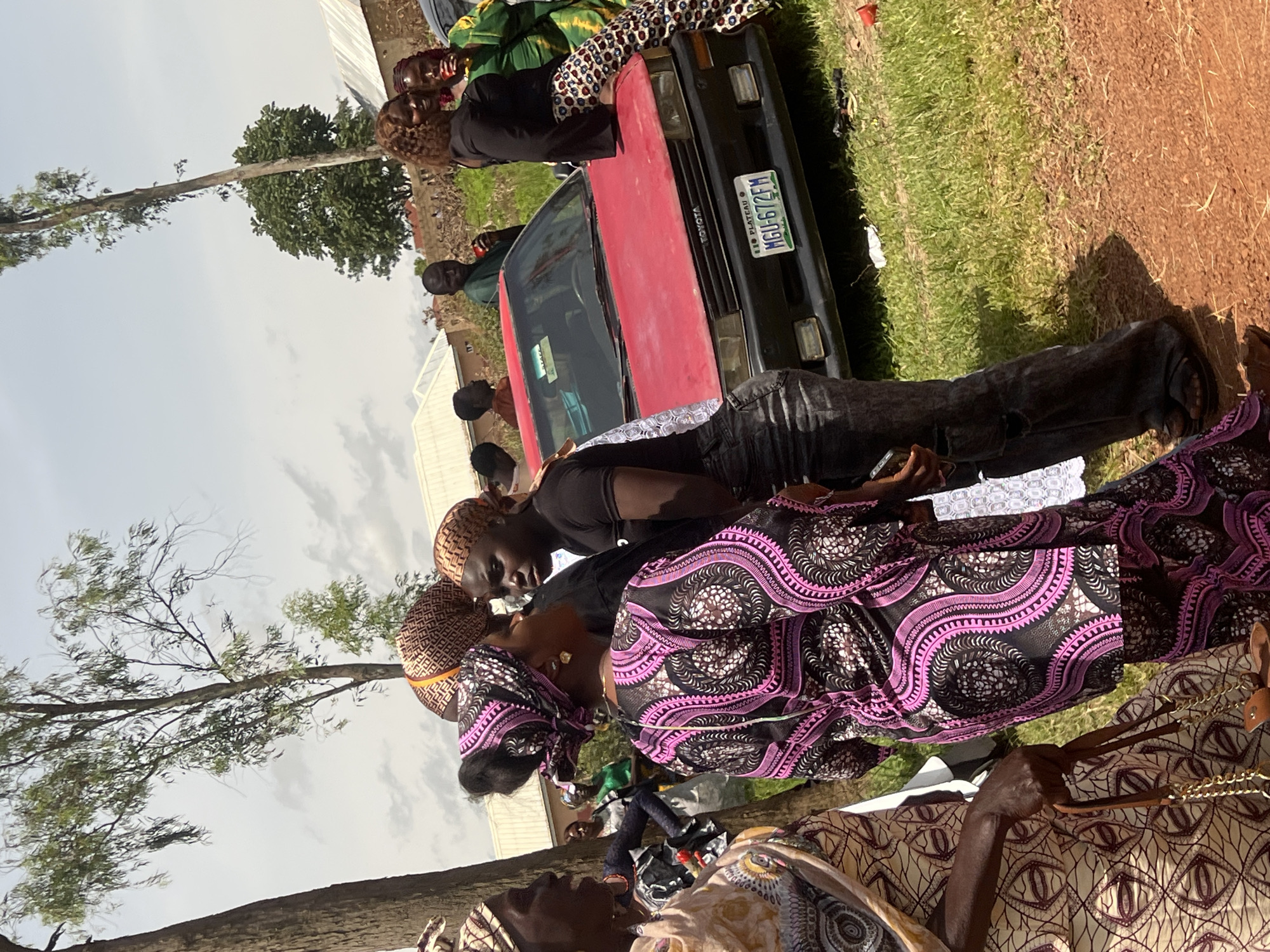 In an outdoor setting in the city of Unknown, Nigeria, a group of people are gathered around a red Toyota sedan. The sky is partly cloudy, and trees with sparse foliage are visible in the background. The ground is a mix of grass and reddish-brown dirt.
Several individuals are the focus of the image. In the foreground, a woman wearing a light-colored, patterned dress and a headscarf is holding a brown purse with a gold chain. To her right, another woman is wearing a vibrant pink and black patterned dress and a matching headscarf. Her gaze is directed towards a young woman in a black t-shirt and distressed denim jeans, who appears to be speaking to her. This young woman has blonde-streaked hair.
Further back, a man in a green shirt is standing near the car, holding a red object. Other individuals are visible in the background, including a woman in a green outfit with a patterned skirt, and several men. The red Toyota has a license plate that reads "MGU 672FM PLATEAU". The car appears to be parked on the dirt and grass area. The overall scene suggests a social gathering or event.