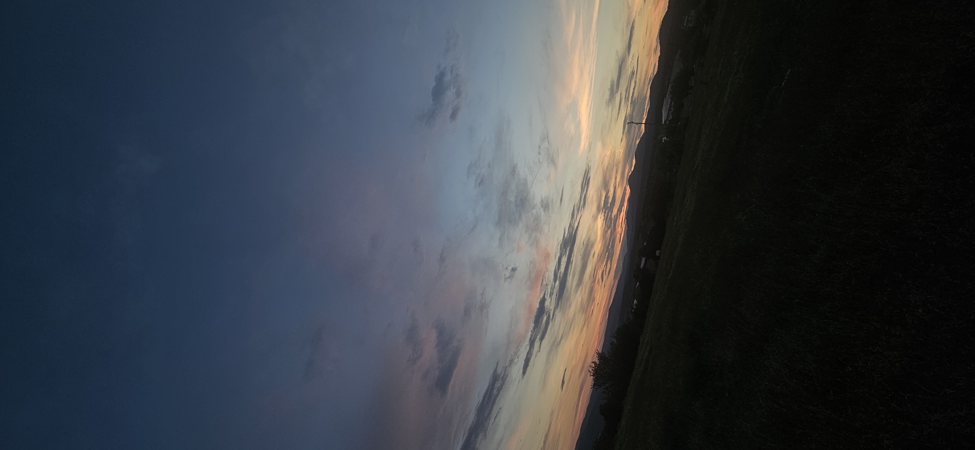 This image depicts a rural landscape at dusk, featuring a rolling hill in the foreground covered in dark vegetation, possibly grass or low-lying crops. The midground reveals a silhouette of distant mountains, with a few faint structures and trees visible against the horizon. The sky dominates the upper portion of the image, transitioning from a deep blue at the top to hues of pink, orange, and light blue near the horizon, indicative of a sunset or twilight. Wispy clouds are scattered across the sky, catching the fading light. The scene appears to be located in Momin Prohod, Bulgaria, given the presence of rolling hills and mountainous terrain characteristic of the region. There are no people or animals visible in the image, nor are any specific actions or interactions taking place. The overall mood is serene and tranquil, capturing the end of the day in a natural setting.