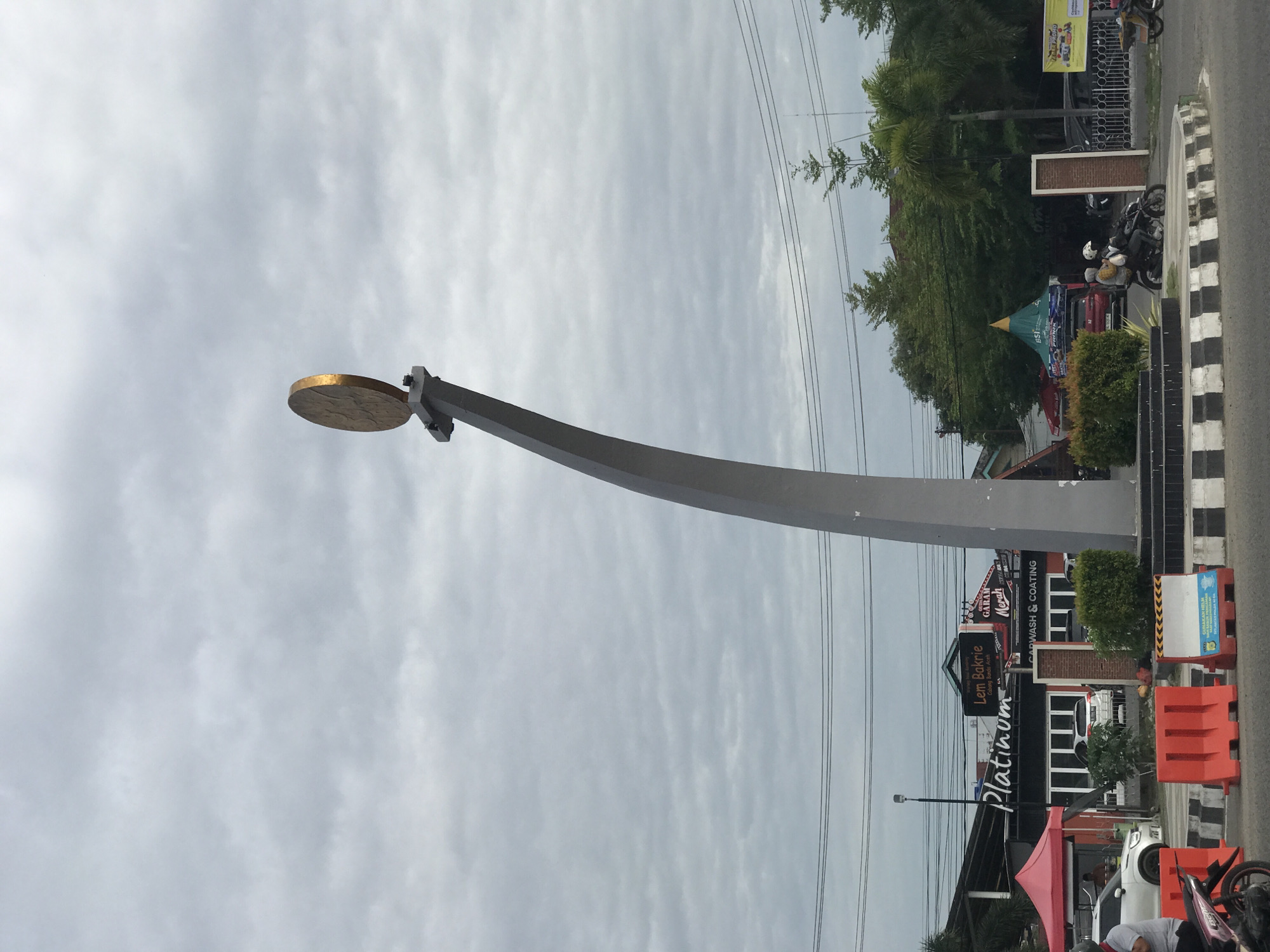 The image depicts a cloudy, overcast day at a roundabout in Lhokseumawe, Indonesia. A large, curved, silver metal sculpture dominates the foreground, topped with a gold disc resembling a coin or medallion. In the background, commercial establishments are visible, including a car wash named "Platinum." Various vehicles, including cars and motorcycles, are parked or moving along the adjacent road. A striped black-and-white curb separates the road from the sidewalk. Trees and other vegetation are also present.