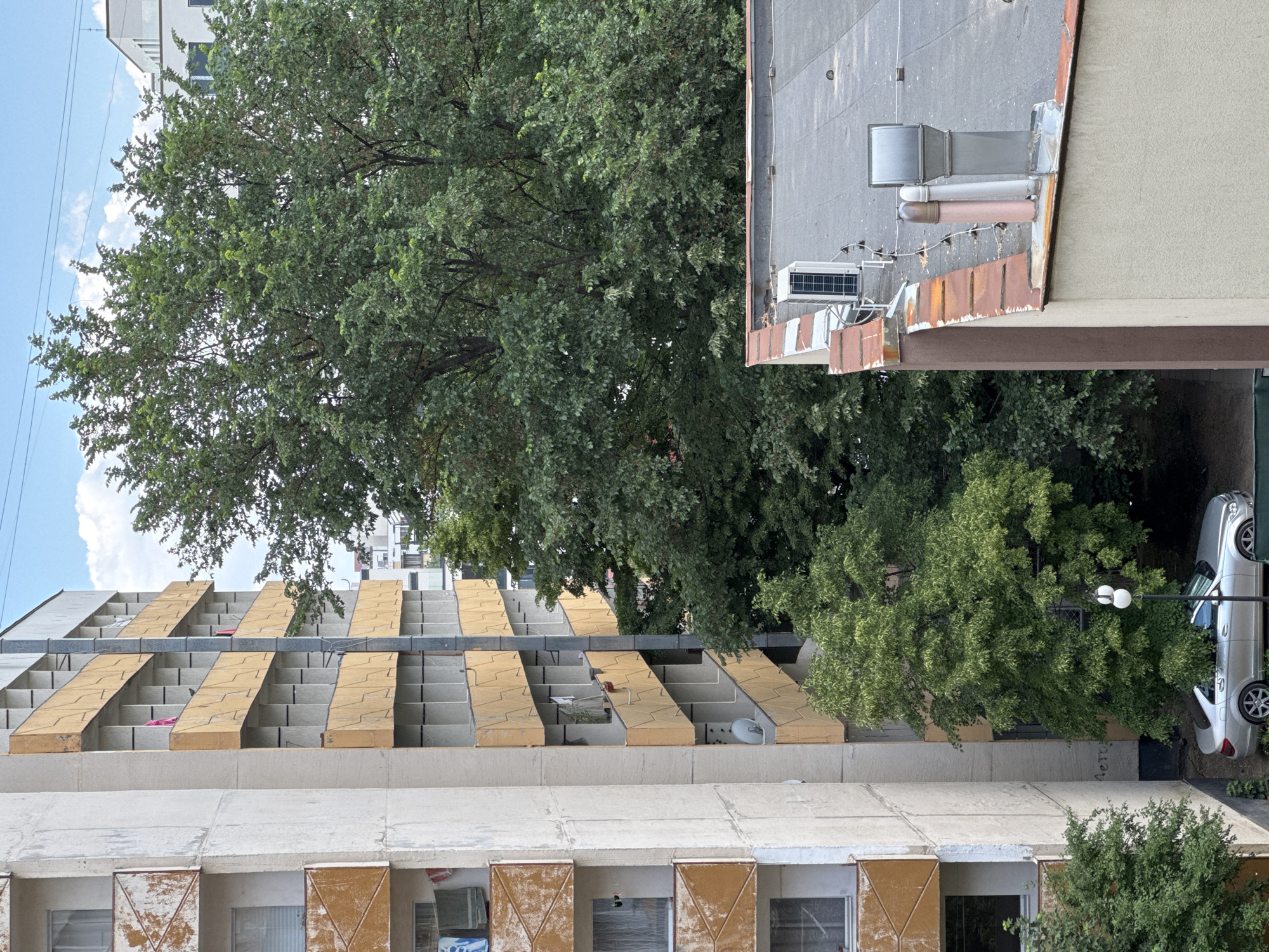 The image depicts a building in Sofia, Bulgaria, viewed from an elevated perspective. The building features a concrete exterior with multiple floors. The balconies are marked by protruding, rectangular structures painted in a faded yellow. Windows on the lower floors are protected by metallic shutters. Adjacent to the building is a densely foliated tree that obscures parts of the structure. To the right of the building, there is a rooftop with mechanical equipment. A silver car is parked in a space between the building and another structure. The sky is visible with light clouds.