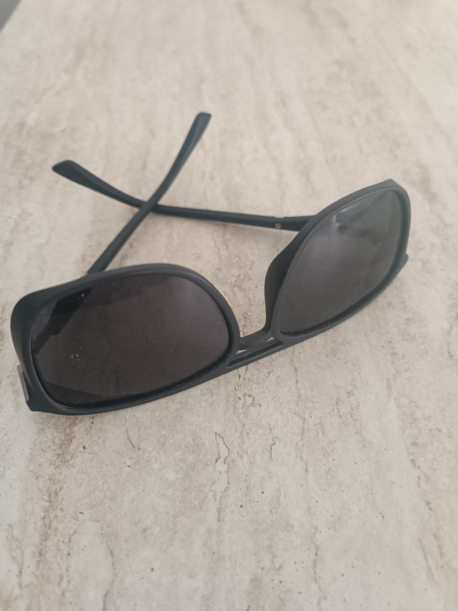 A pair of black sunglasses lies on a beige, speckled surface. The sunglasses are open and oriented diagonally across the frame, with the lenses facing upwards. The surface appears to be a textured stone or countertop. There are no people or other objects visible in the image. The location is unknown.
