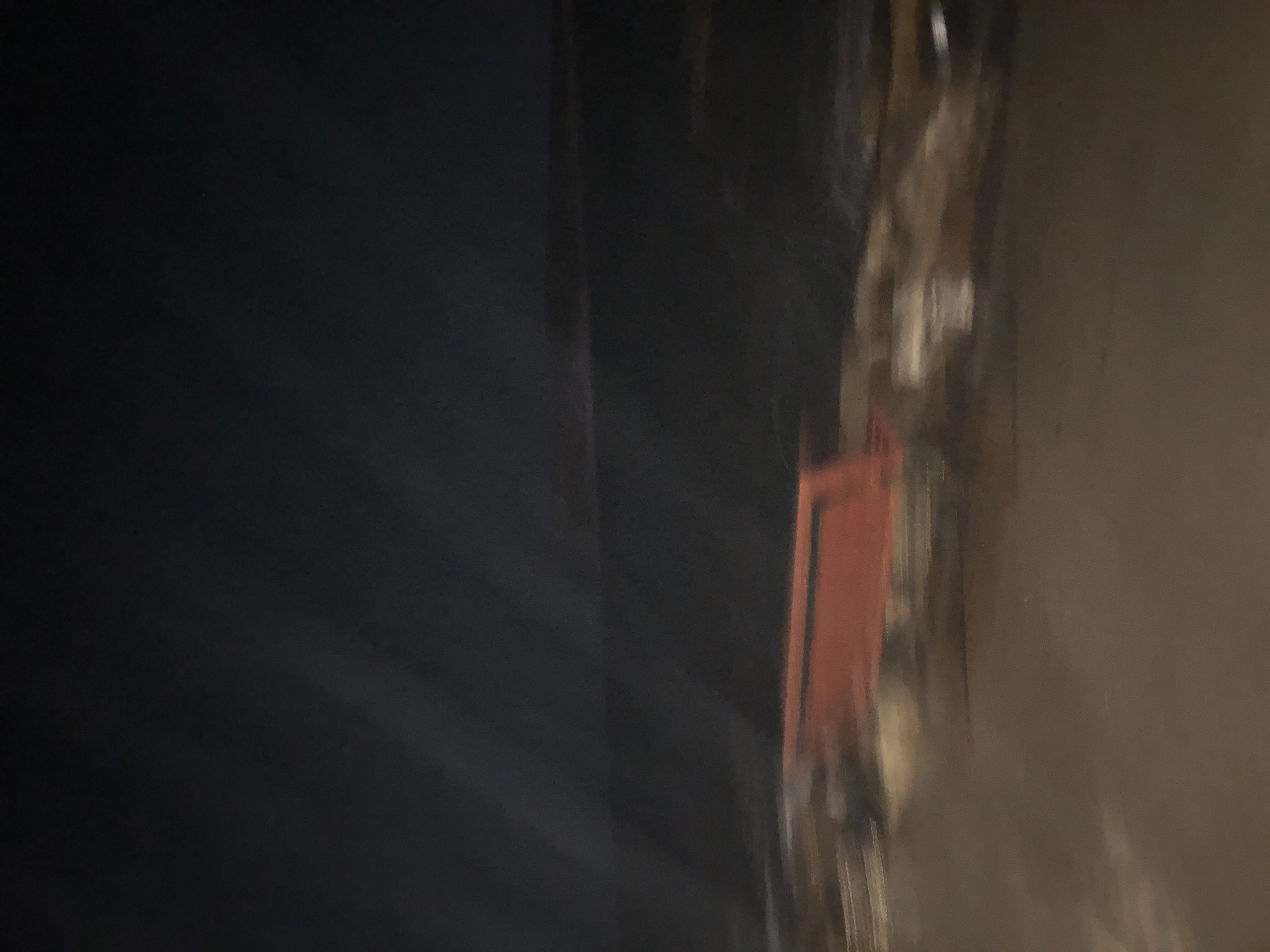 The image shows an out-of-focus view of an outdoor scene at night, likely in Karaburun, Turkey. A red bench is visible in the foreground, placed on a paved surface. Behind the bench, a body of water stretches to the horizon under a dark sky. The water is dark and indistinct, suggesting it's nighttime. Streaks of light are present in the upper left portion of the image, possibly from artificial lighting or a light source moving across the frame. The overall impression is of a dimly lit, serene, and possibly deserted location.