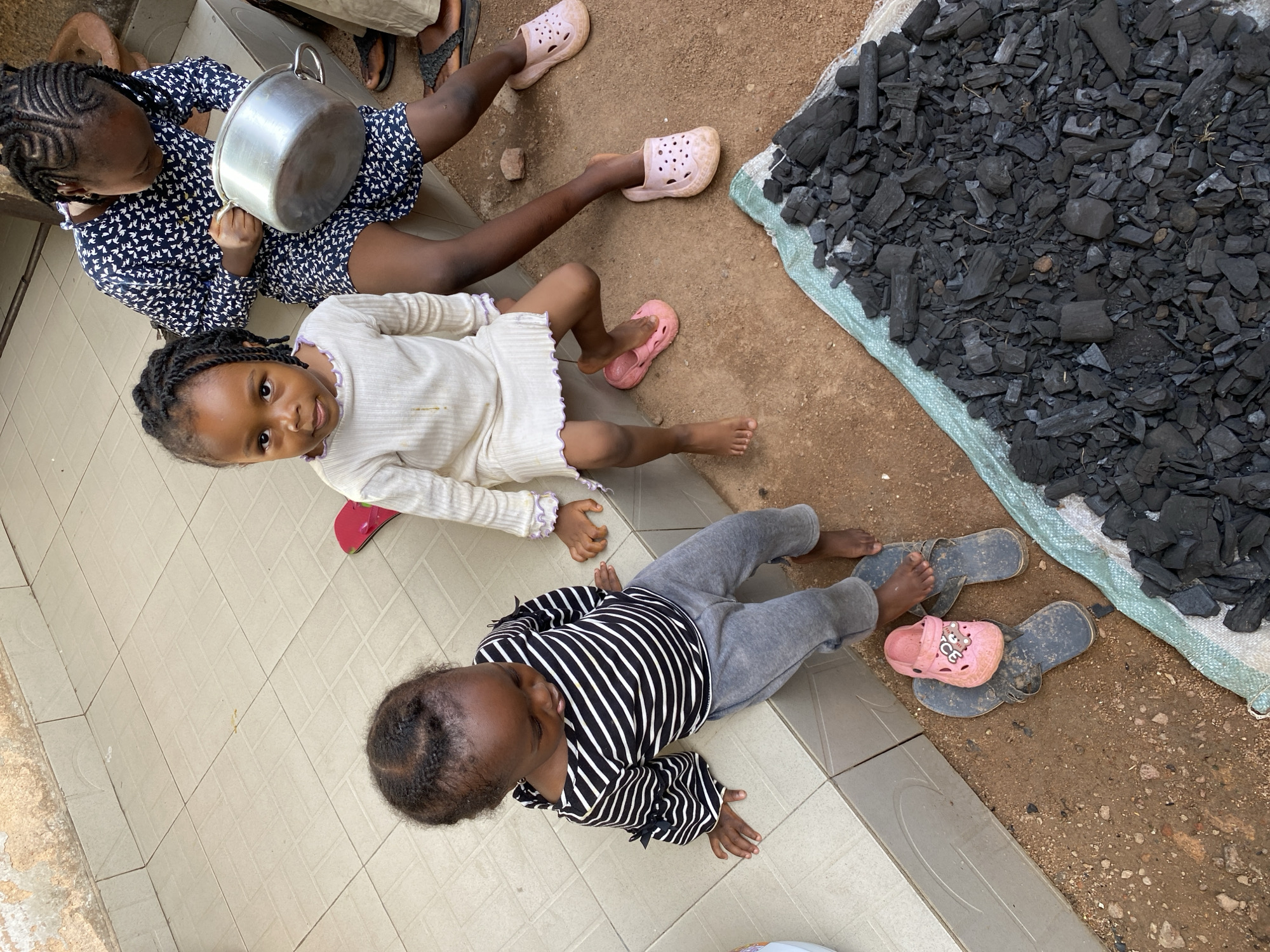 Three young African girls sit on the ground in Bukuru, Nigeria. One girl, wearing a white dress, looks directly at the camera. Another girl holds a metal pot. The third girl wears a striped shirt. A pile of charcoal is near the girls. Two of the girls wear pink footwear. Two pairs of adult sandals and a child's pink sandal lie near the charcoal. The ground surface varies from tiles to dirt.