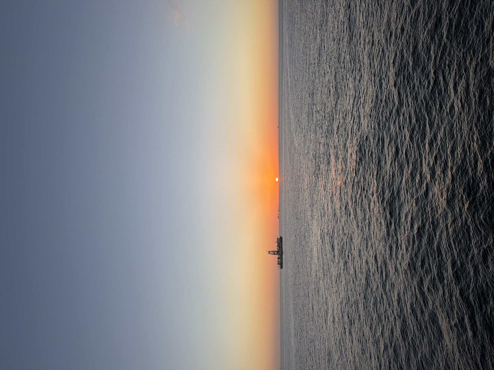 The image captures a vast seascape at either sunrise or sunset, dominated by a striking horizon line. The sky transitions from a soft, hazy blue-grey on the left to warm oranges and yellows around the sun, which appears as a bright, circular orange orb positioned low on the horizon, slightly above the water line, towards the right side of the frame.
The water surface, covering the bottom half of the image, is dark and textured with numerous small ripples and waves, reflecting the low light and casting subtle shadows that give it a metallic appearance. The light from the sun creates a shimmering path across the water extending towards the viewer.
Silhouetted against the vibrant sky and distant horizon is a large, industrial-looking structure or vessel, possibly an offshore oil rig or a cargo ship with a prominent superstructure, situated slightly to the left of the sun. Its intricate shape is clearly visible despite being a silhouette. Further along the horizon, both to the left and right of the main structure, smaller, fainter silhouettes of other vessels or offshore installations can be discerned, indicating a maritime setting with active industry or shipping traffic.
The overall mood is serene and contemplative, highlighting the beauty of the natural phenomenon of the rising or setting sun against an industrial backdrop at sea. There is no visible text in the image.