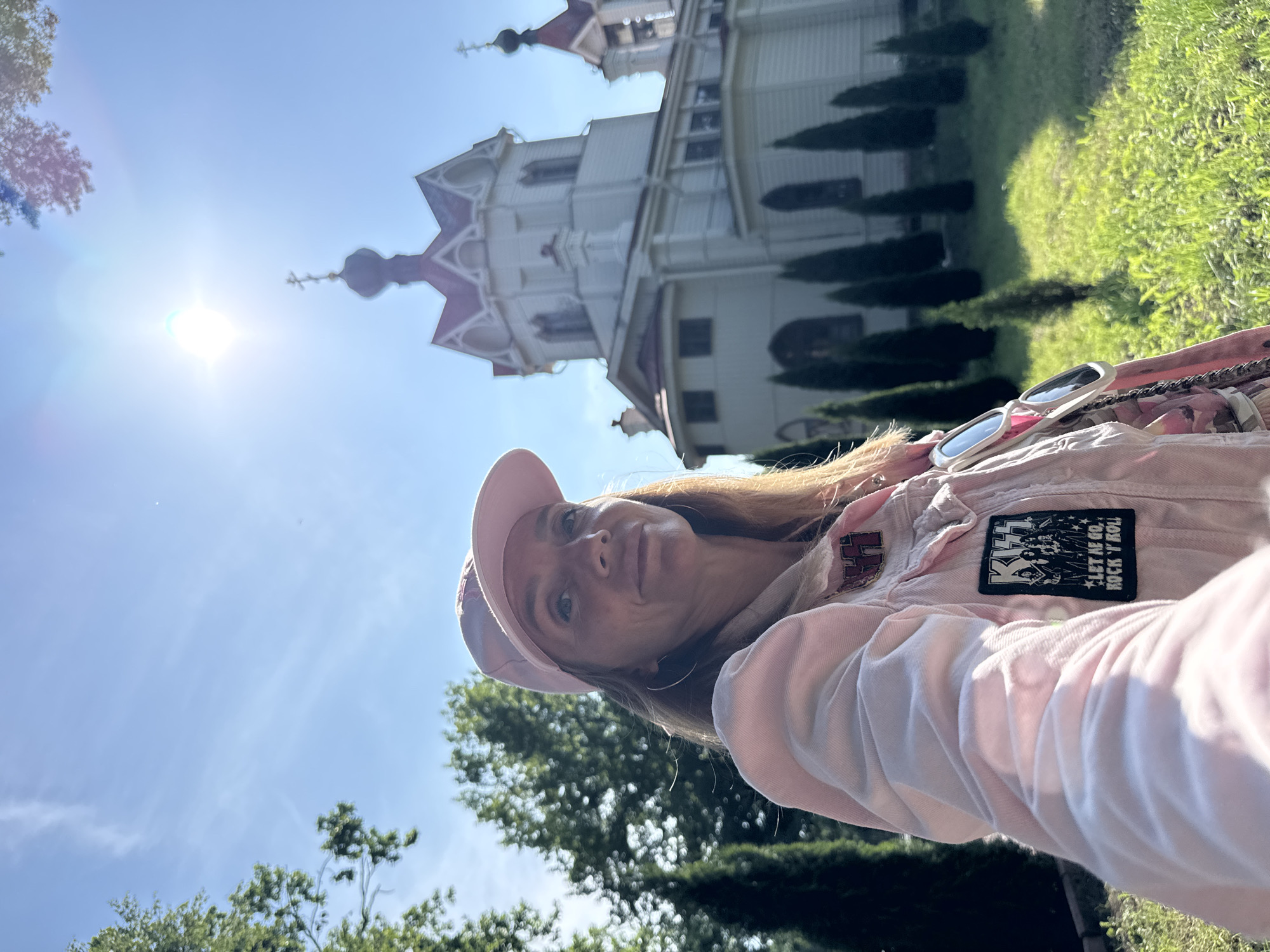 A woman wearing a pink baseball cap and a light pink jacket with patches stands outside a white and pink building with ornate architectural details, possibly a church. The building has multiple spires and decorative elements. To the right of the building, a lush green grassy area and dark green trees are visible. The sun is shining brightly, creating lens flare and casting shadows. The woman is looking towards the camera, with a slight smile. She is wearing hoop earrings and sunglasses are resting on her jacket. One of the patches on her jacket is a black rectangle with the KISS logo and text that reads "LET ME GO. ROCK 'N' ROLL". Another patch is red and silver with the KISS logo. The image appears to be taken on a sunny day. The overall scene suggests an outdoor excursion on a clear day in a location with a visually striking building.
