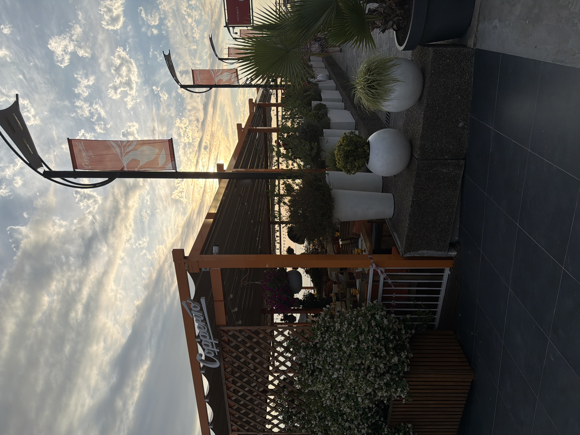 This is a medium-shot, eye-level, outdoor, daytime photograph of a restaurant with a prominent sign reading "Copperto". The restaurant appears to be located in Sochi, Russia, as indicated by the banner on a lamppost which reads "Novikov". The scene is set during sunset, with a cloudy sky illuminated by the setting sun. The restaurant has an orange awning and a wooden lattice section. Several tables and chairs are visible through the open-air seating area. In the foreground, there are various potted plants, including a large shrub with white flowers on the left and several white spherical planters containing greenery. A palm tree is visible on the right side of the image. The ground is paved with dark square tiles. A person is walking in the background on the right side of the image. The overall atmosphere is calm and serene, with the warm tones of the sunset casting a gentle light on the scene.