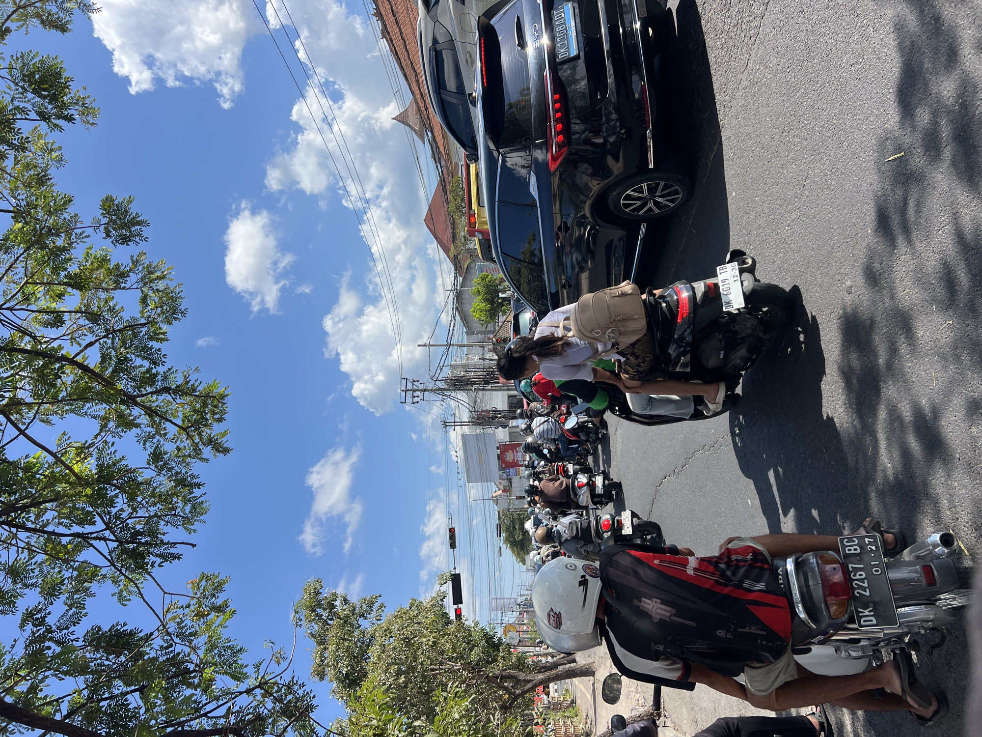 The image captures a traffic jam on a sunny day in Denpasar, Indonesia, likely on a main road. In the foreground, a person wearing a white helmet and a patterned shirt rides a motorcycle, with another motorcycle visible behind it. The road is filled with numerous other motorcycles, some carrying passengers, creating a dense flow of traffic. A black SUV is prominent in the middle ground, with a license plate clearly visible. In the background, buildings and utility poles suggest an urban environment. The sky is bright blue with scattered white clouds, and trees with green foliage frame the top of the image. The presence of multiple vehicles and the traffic signal indicates a busy intersection.
