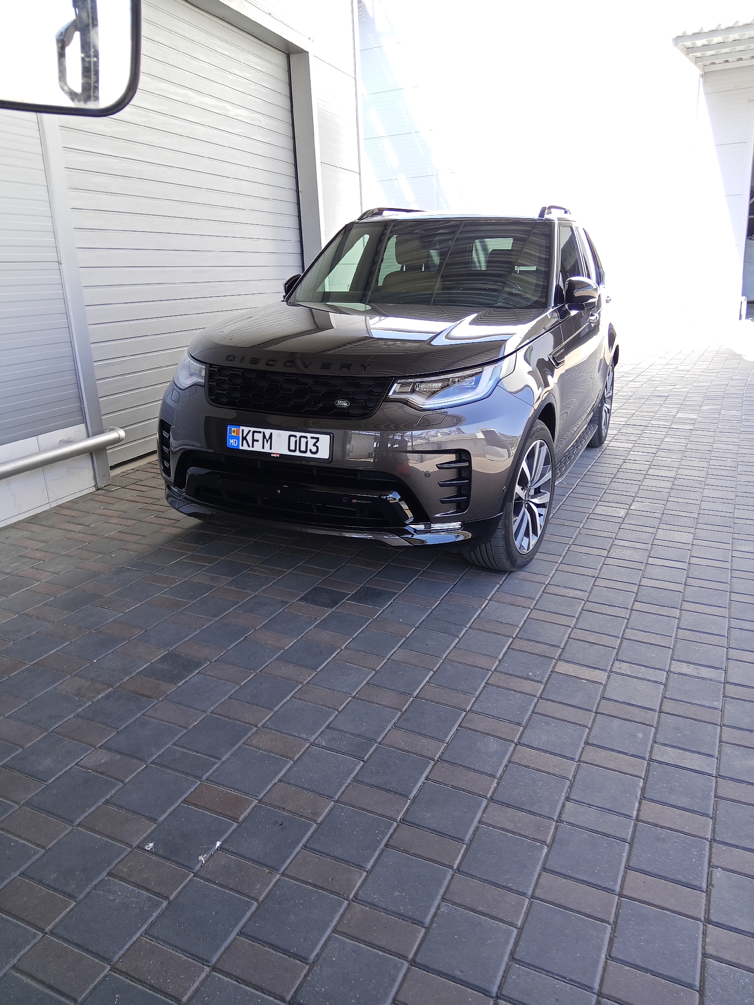 An eye-level, medium shot captures a dark gray Land Rover Discovery parked on a patterned gray and dark brown paved driveway in front of a white corrugated metal garage door in the city of Ceadîr-Lunga, Moldova. The vehicle's front end, including its headlights, grille with "DISCOVERY" lettering, and a Moldovan license plate reading "KFM 003," is prominently displayed. The car's reflection is visible on its polished surface, and the sunlight creates bright reflections on the garage door and the upper right portion of the image. A side mirror of another vehicle is partially visible in the upper left corner. The scene appears to be a commercial or residential garage setting.