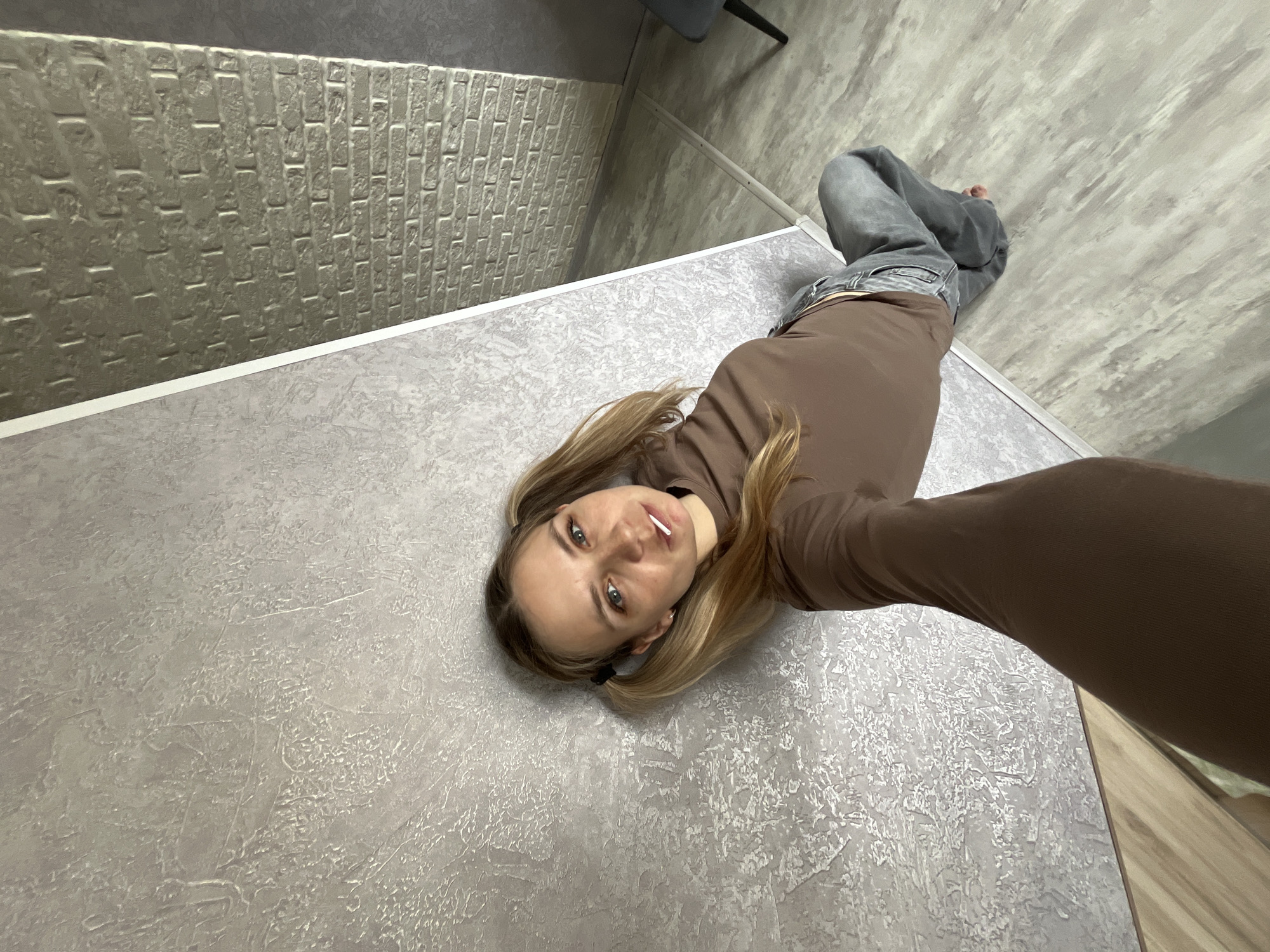 An interior wide-angle photograph, taken in a residence in Chaikovsky, Russia, depicts an adult female capturing a selfie. The woman, with fair skin, light eyes, and blonde-to-light-brown hair styled in two pigtails, wears a brown long-sleeved top and loose grey jeans, and is barefoot. A white lollipop stick is visible between her lips. She maintains direct eye contact with the camera, which is held by her extended right arm, partially visible in the lower foreground. The background on the left features a light grey wall with a coarse, irregular textured finish. This wall transitions into a lighter grey brick-patterned section via a narrow white vertical trim. A darker grey wall is visible beyond the brickwork on the right. The floor is light grey laminate or vinyl, displaying a subtle, cloudy pattern. A dark chair leg is partially visible in the bottom right corner.