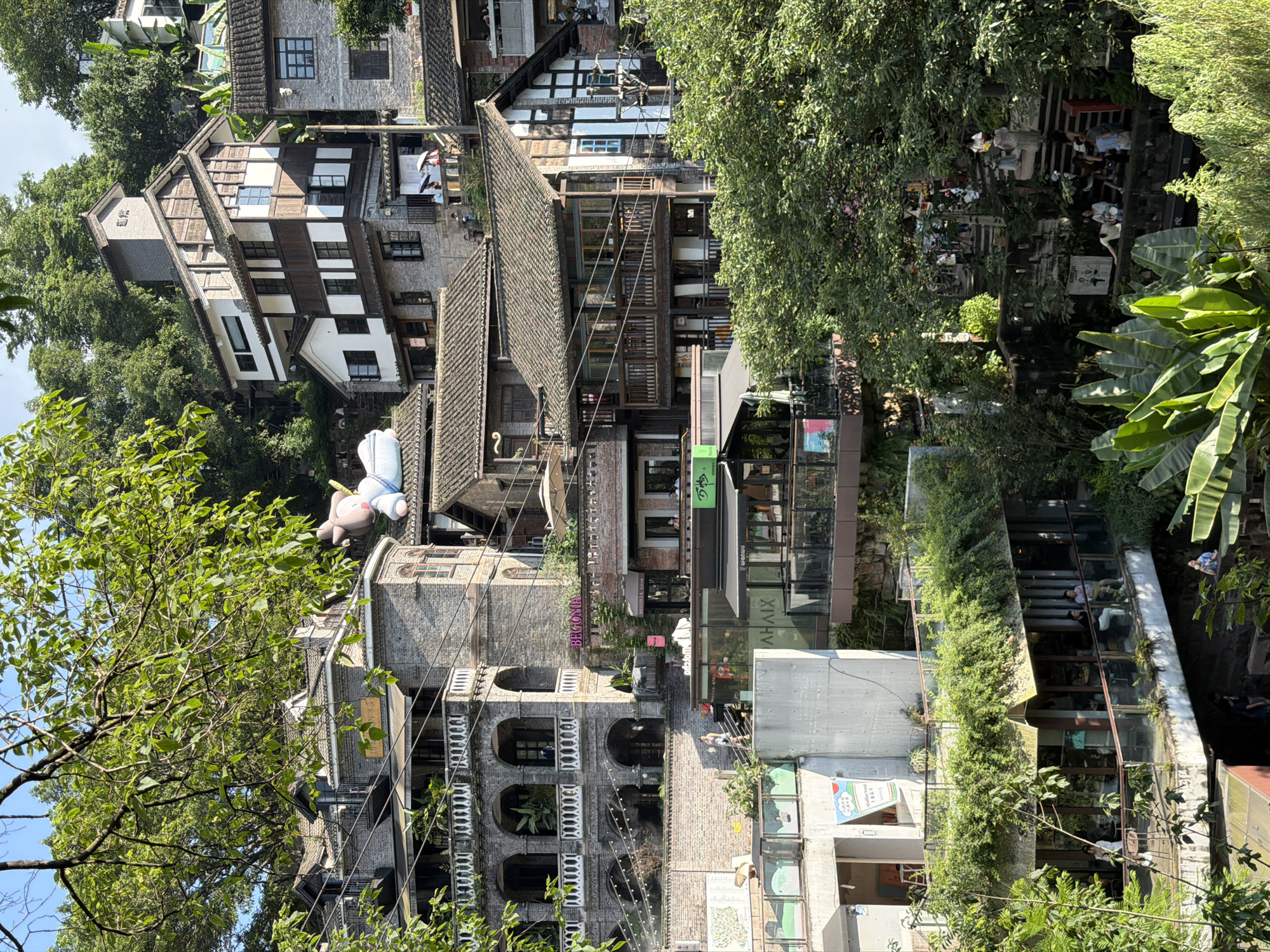 The image depicts a vibrant, multi-layered architectural scene in Nan'an District, China, characterized by buildings constructed on a steep hillside amidst lush greenery. The architecture is a blend of traditional Chinese and modern styles, with many buildings featuring dark tiled roofs, exposed wooden beams, and stone facades. Several levels of walkways, balconies, and staircases connect the buildings, creating a complex and visually interesting urban landscape.
A prominent element in the upper portion of the image is a large, inflatable cartoon character, possibly a pig, resting on a rooftop. In the mid-ground, a building with arched windows and balconies, adorned with potted plants, stands out. Below this, more modern structures with glass facades and balconies house shops and cafes. People are visible on various levels, some walking, some seated, suggesting a lively public space. The environment is rich with vegetation, including large trees with dense foliage, vines trailing down walls, and large tropical-looking leaves in the foreground, indicating a humid, perhaps sub-tropical climate. The overall impression is one of a bustling, architecturally unique district integrated with nature.