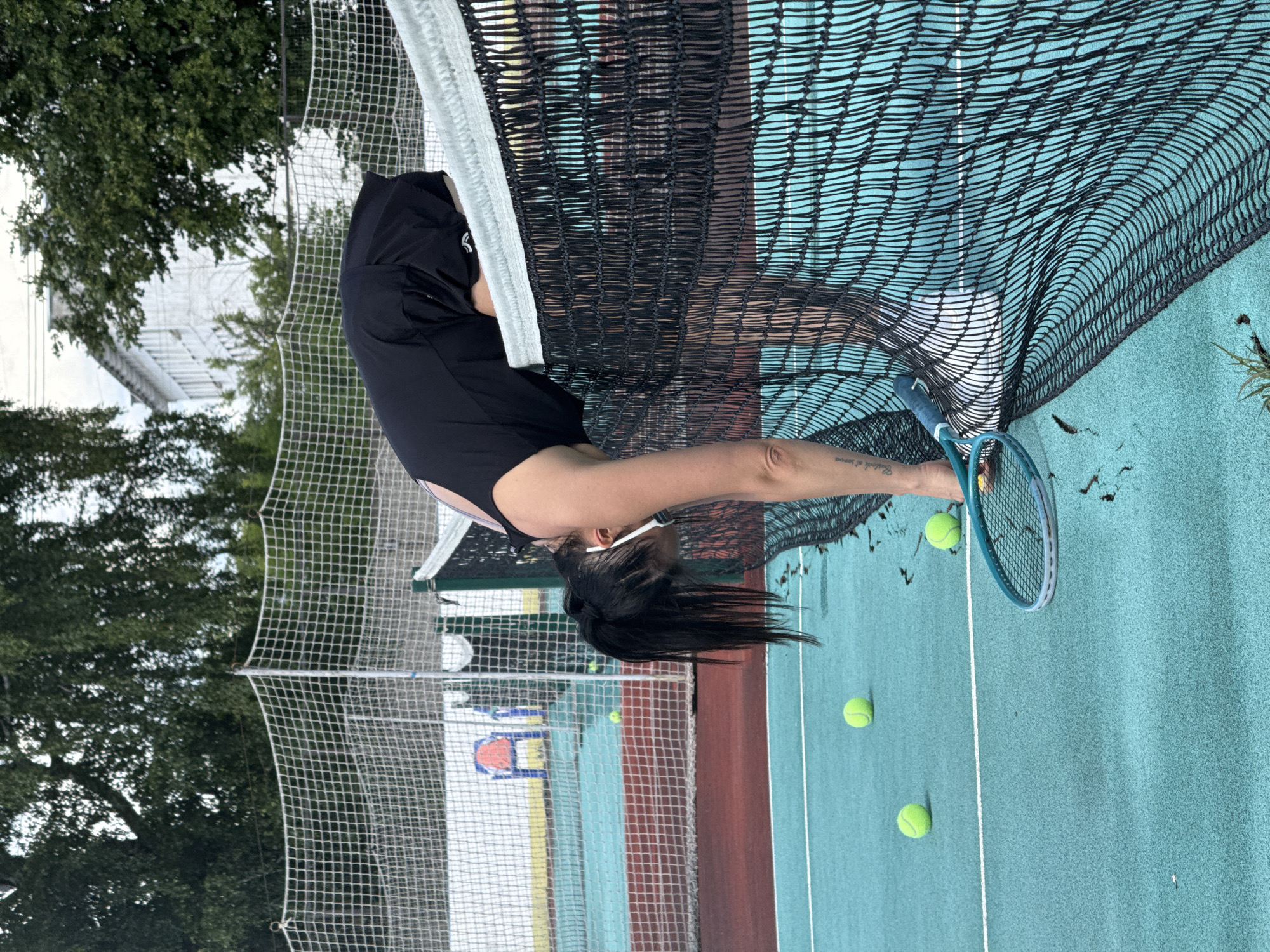 A woman with dark hair tied back in a ponytail is on a tennis court, bent over and reaching for a tennis racket. She is wearing a black athletic outfit. Tennis balls are scattered on the teal-colored court. The scene is outdoors, with trees and a white structure visible in the background, along with a tennis net and fence. The sky appears overcast, suggesting a cloudy day. The woman's pose suggests she is either retrieving a dropped racket or preparing to hit a low shot. There is visible text on the woman's forearm, a tattoo that reads "Rest in peace." The image orientation is rotated 90 degrees clockwise.