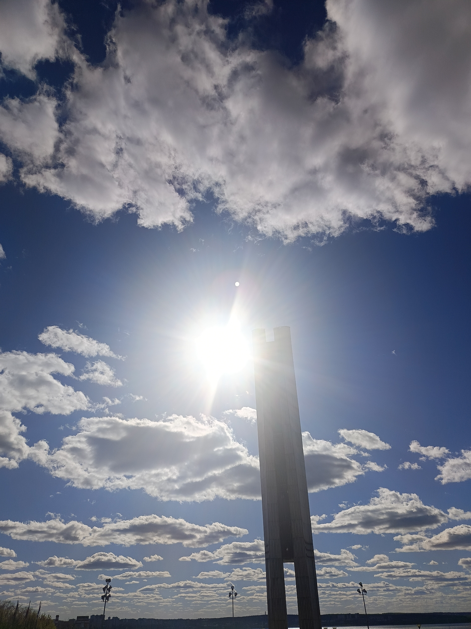 The image captures a bright, sunny day in Izhevsk, Russia, showcasing the city's iconic monument, the Monument to the Fighters of the Revolution. The monument, a tall, slender, concrete structure, dominates the foreground. The sun is positioned directly behind it, creating a strong solar flare effect that obscures some details. The sky is a vibrant blue, scattered with numerous fluffy, white cumulus clouds. In the distance, a cityscape is visible, including a body of water and lampposts. The overall scene is a wide-angle shot, emphasizing the monument's height against the expansive sky.