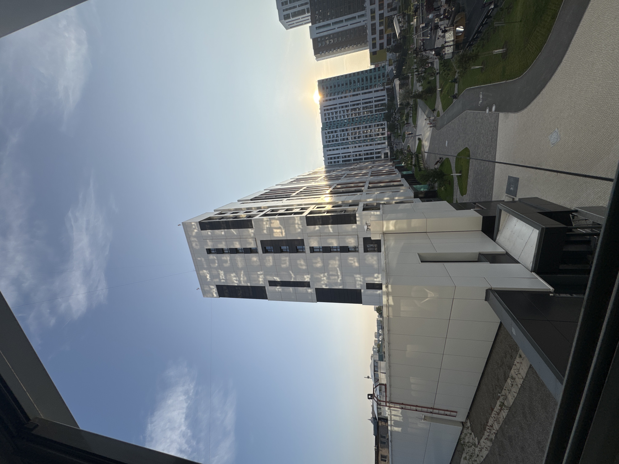 The image captures a modern urban residential complex bathed in the warm glow of a setting sun. In the foreground, a contemporary building with a white facade and dark geometric accents dominates the frame. Its reflective surface mirrors the sky and surrounding structures. To the left, a lower white building with a red ladder attached to its side is visible. Further into the scene, a cluster of multi-story apartment buildings stretches across the horizon. One building in the mid-ground features a striking turquoise and white facade, while others exhibit a mix of white and darker tones. The setting sun is visible between two of these buildings, casting a warm light and creating lens flare. A paved walkway winds through a landscaped courtyard area with patches of green grass and trees. Several people are scattered throughout the courtyard, some walking and others gathered near playground equipment. The sky above is a clear blue with wispy white clouds, and a thin wire is suspended across it. The scene appears to be part of a new development, possibly in a suburban or newly urbanized area of Novosibirsk, Russia.