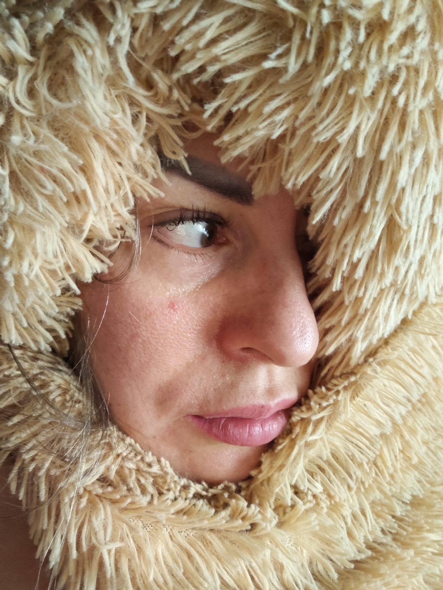 A close-up, profile shot of a woman's face is captured, looking to the left. She is partially obscured by a fluffy, tan-colored scarf or hood. Her left eye is visible, with dark eyelashes and a light reflection on the iris. A small, red blemish is noticeable on her left cheek. Her nose is prominent, and her lips are slightly parted, revealing a pinkish hue. Strands of grey-streaked brown hair are visible. The background is out of focus, with a warm, neutral tone. The image is undated and location-unspecified, though the prompt suggests it is in Almaty, Kazakhstan.