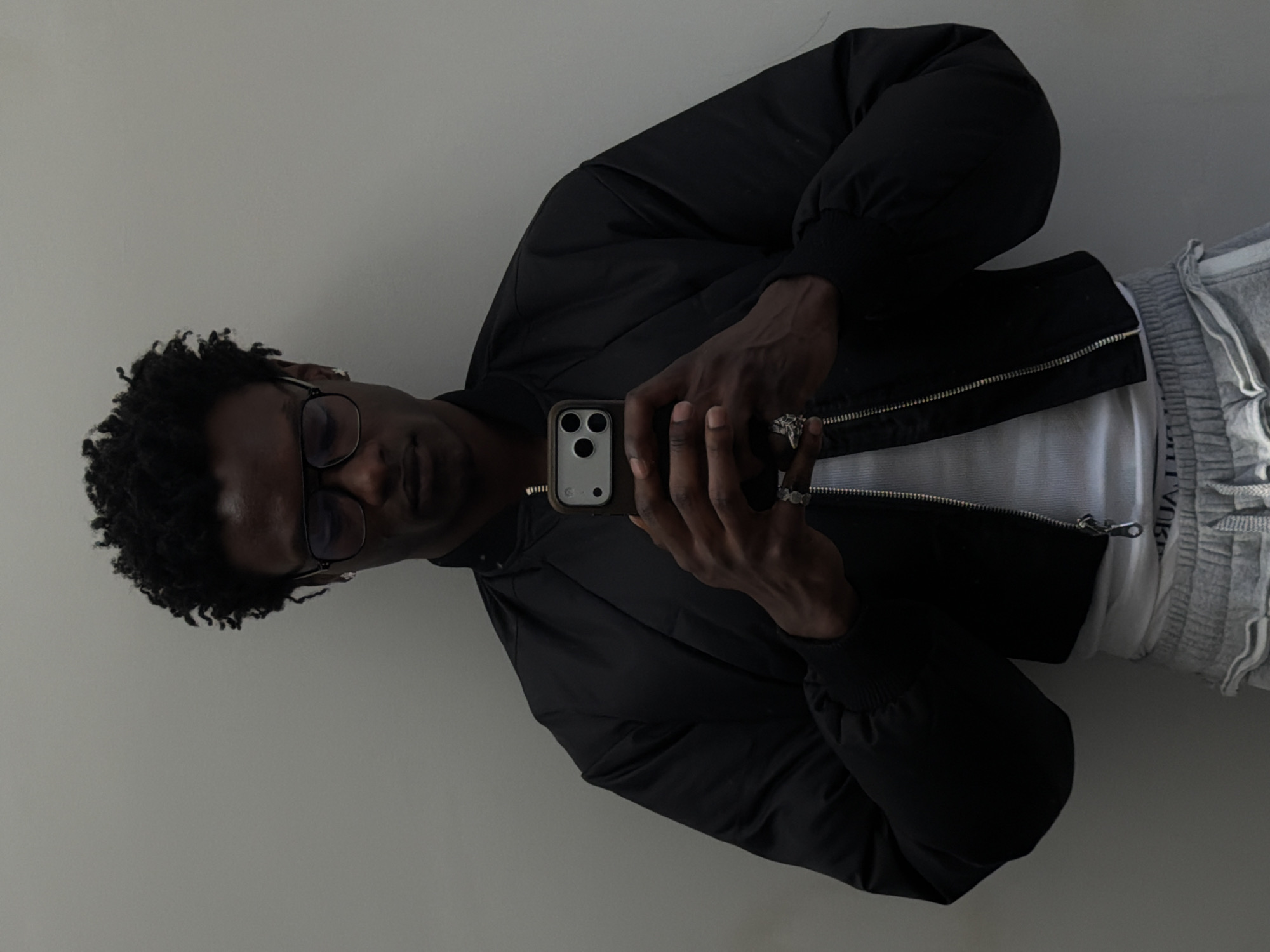 A person is holding a smartphone and taking a selfie. The person is wearing glasses, a black jacket, a white shirt, and gray sweatpants. The background is a plain white wall. The location is not discernible from the image.