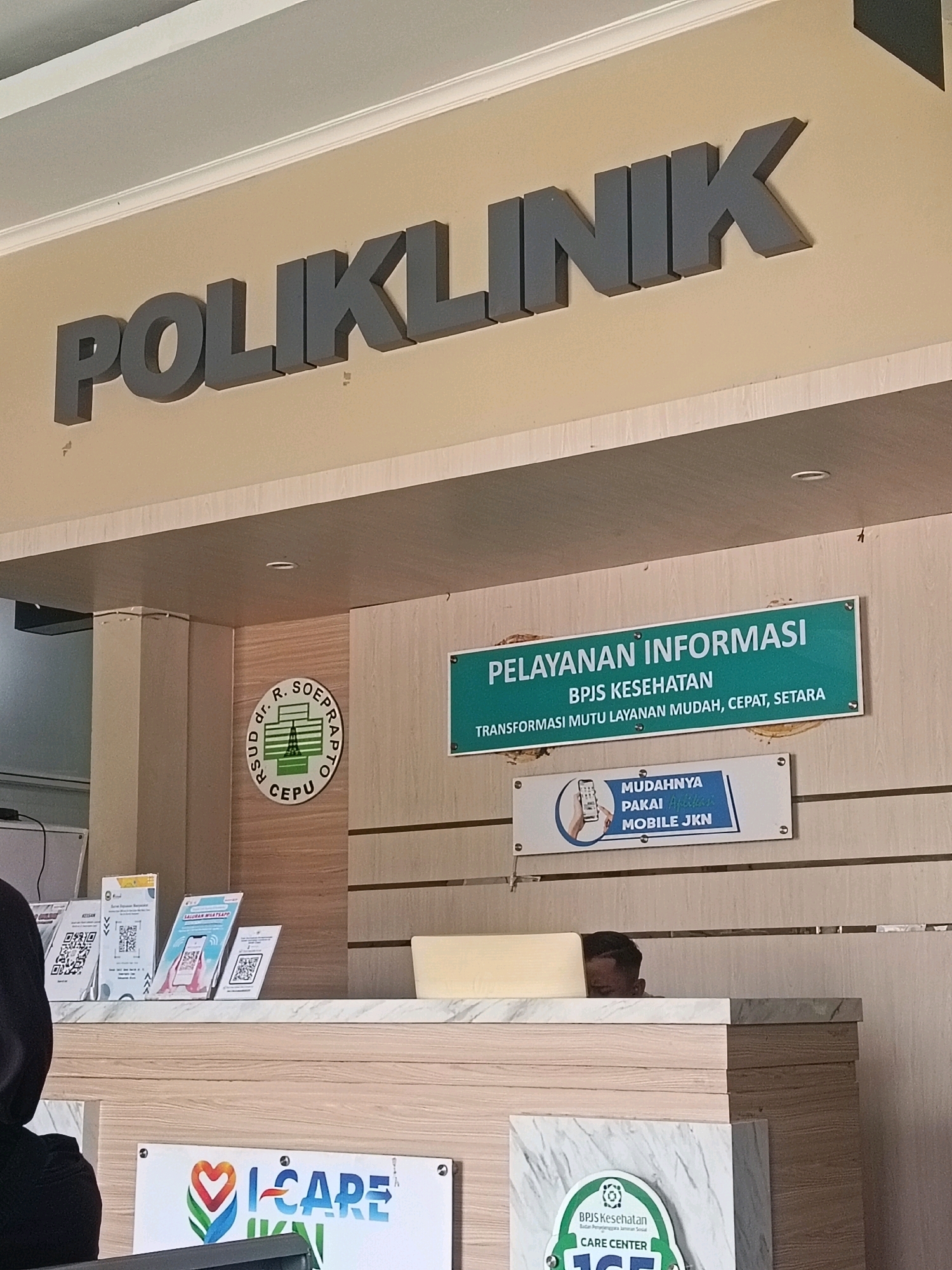 A medium shot captures a hospital's reception area in Balun, Indonesia. A prominent sign above reads "POLIKLINIK" in large, dark grey 3D letters. To the left, a circular emblem is visible, with text that appears to be "RSUD DR. R. SOEPRAPTO CEPu". To the right of the emblem, a green sign displays "PELAYANAN INFORMASI BPJS KESEHATAN TRANSFORMASI MUTU LAYANAN MUDAH, CEPAT, SETARA" in white text. Below this, a smaller sign shows an illustration of a hand holding a smartphone, with text that reads "MUDAHNYA PAKAI MOBILE JKN". Several QR codes are displayed on the counter to the left. A person, partially visible, is seated behind the counter, looking down at a laptop. The reception counter is made of wood and marble. In the lower portion of the image, signs with the text "I-CARE" and "BPJS Kesehatan CARE CENTER" are visible.