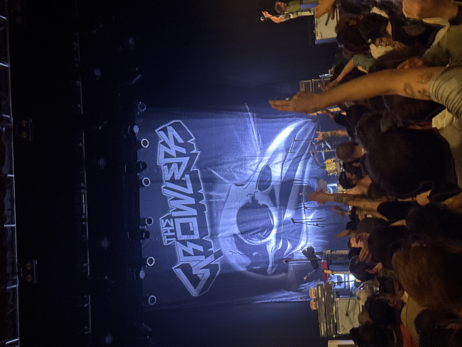 This image depicts an indoor live music concert scene. The event is located in Paris, France. The central feature is a stage dominated by a large backdrop banner displaying "THE GROWLERS" in stylized, block letters above a detailed graphic of a raven or crow skull. The stage is illuminated by spotlights from above. On the left side of the stage, a keyboard setup with amplifiers underneath is visible, topped by a plate holding two cheeseburgers. Multiple microphone stands are present across the stage. To the right, a drum kit with metallic, possibly gold-colored, drums is set up. A male performer with long hair and a beard, wearing a light-colored shirt, stands on the far right of the stage, holding a microphone and gesturing to the audience. Another performer is partially obscured behind the keyboard on the left. The foreground is filled with a dense crowd of attendees, facing the stage. Many individuals have their hands raised in the air; some display a "rock on" hand gesture, indicating active participation and engagement with the performance.