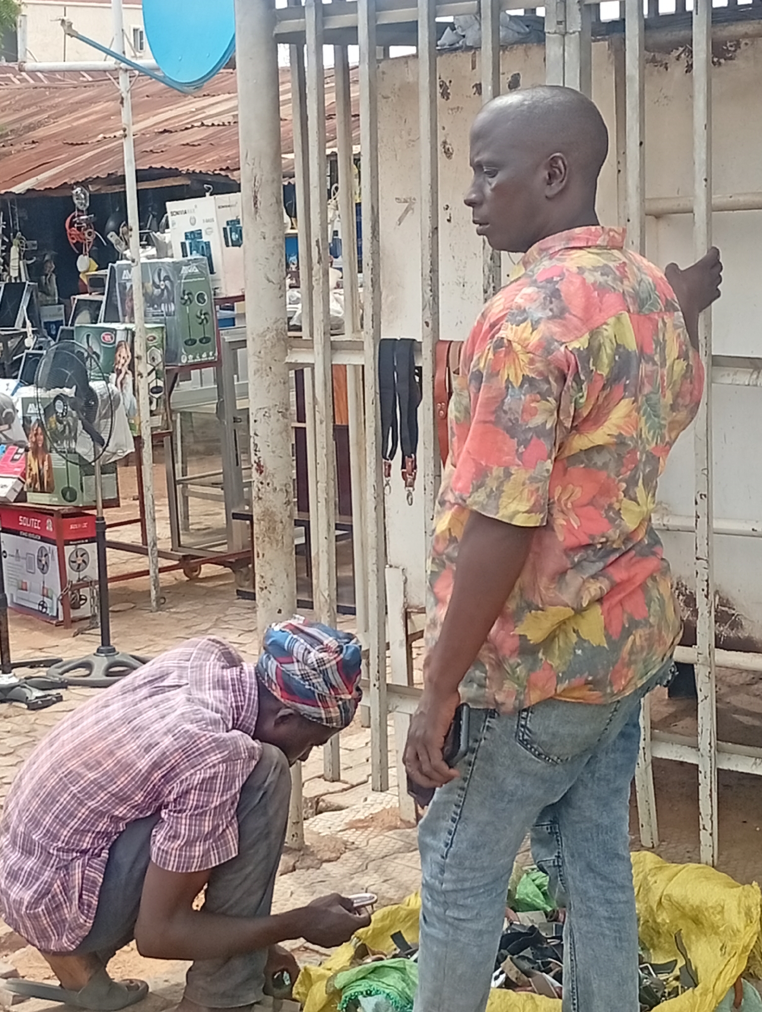The image shows two men in an outdoor setting in Sabon Tasha, Nigeria. One man is squatting, examining something on the ground. He wears a checkered shirt and a head wrap. The other man is standing, facing right, and holding onto a white metal gate. He has short hair and is wearing a colorful shirt and jeans. Bags and metal objects are on the ground. In the background are items for sale, including boxes and fans. A metal structure with a blue satellite dish is visible above. The environment appears to be a commercial area.