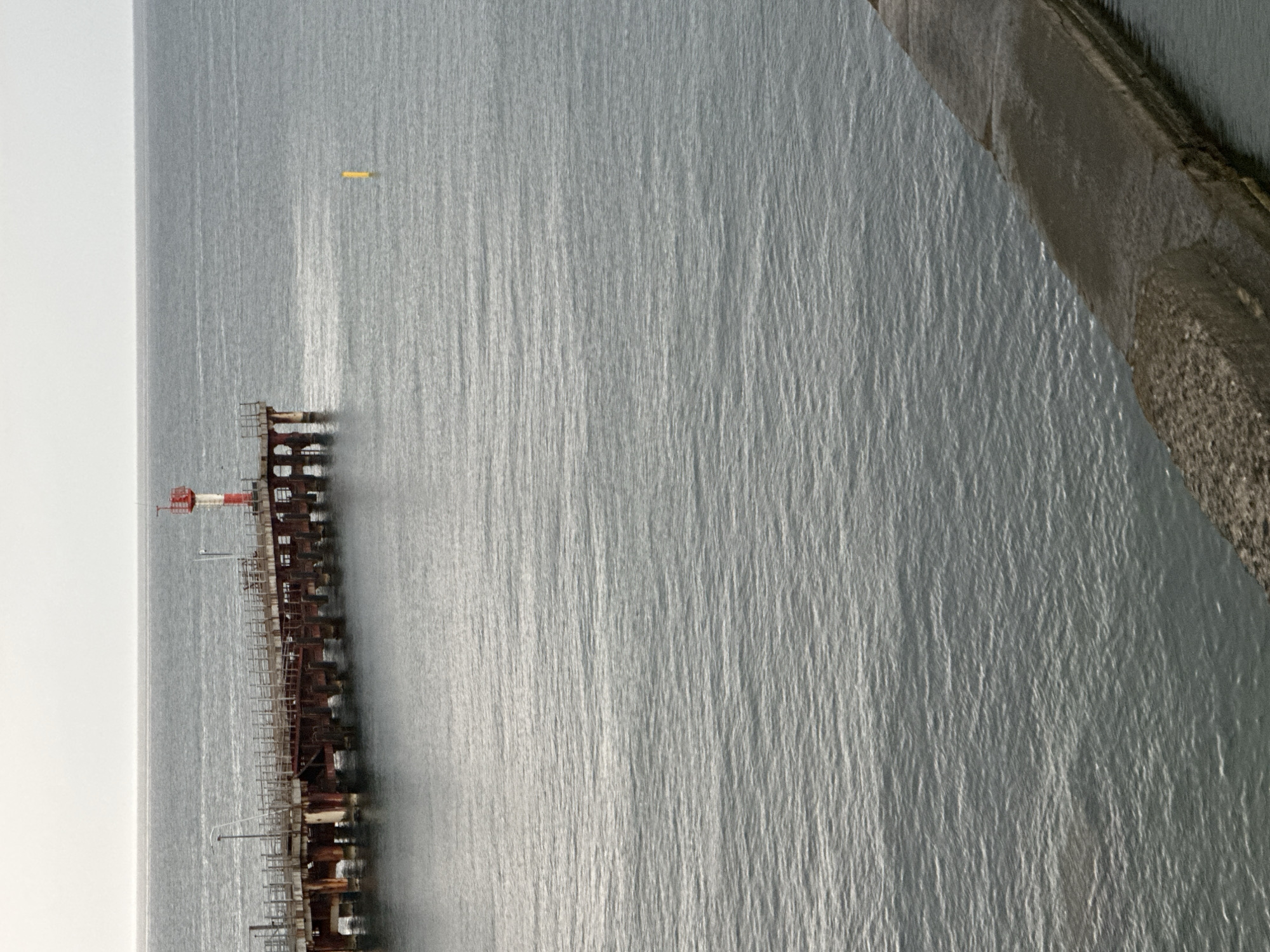 An exterior, medium shot captures a weathered pier extending into a calm, grey sea under an overcast sky. The pier is constructed of dark, rusted metal supports and a wooden walkway. A red and white lighthouse stands at the end of the pier. A small, yellow buoy floats in the water to the right of the pier. In the foreground, a concrete breakwater juts into the frame from the right, its textured surface partially submerged. The water is choppy with a slight sheen reflecting the sky. The location is unknown.