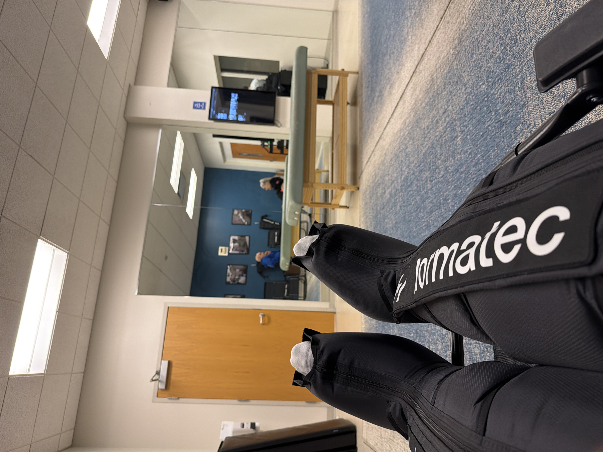The image shows a first-person perspective of a person wearing black compression boots on their legs, with light gray socks visible at the top. The boots have zippers and a white logo that reads "Normatec". The person is seated in a room that appears to be a physical therapy or recovery space. The room has a tiled ceiling with fluorescent lighting, and beige walls. To the left, there is a closed wooden door. In the background, there is a large mirror reflecting a section of the room. The mirrored reflection shows two massage tables with light green padding, and a wooden frame. Behind the tables, there are two people, one sitting and one standing, their faces are not clearly visible. Further back in the reflection, there is a wall with framed black and white photographs. On the right side of the room, a black flat-screen monitor is mounted on a white column. The floor is carpeted with a blue and white pattern. The overall setting suggests a professional facility. Based on the context clues within the image, this scene is likely located in Beachwood, Ohio, United States of America, potentially within a sports medicine clinic or a rehabilitation center.
