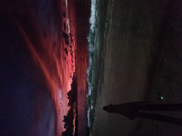 The image depicts a beach scene at dusk in Pecatu, Indonesia. The sky transitions from dark purple and blue overhead to vibrant reds and oranges along the horizon, marked by dark, scattered clouds and two prominent diagonal red streaks in the upper atmosphere. The ocean, appearing dark, exhibits white-capped waves breaking and rolling towards the shore. A distant, silhouetted landmass with faint lights is visible on the left horizon. In the lower-left foreground, a solitary human figure stands in silhouette on the dark sand, facing the water. A small, bright green glowing object is present on the sand near the figure's feet.