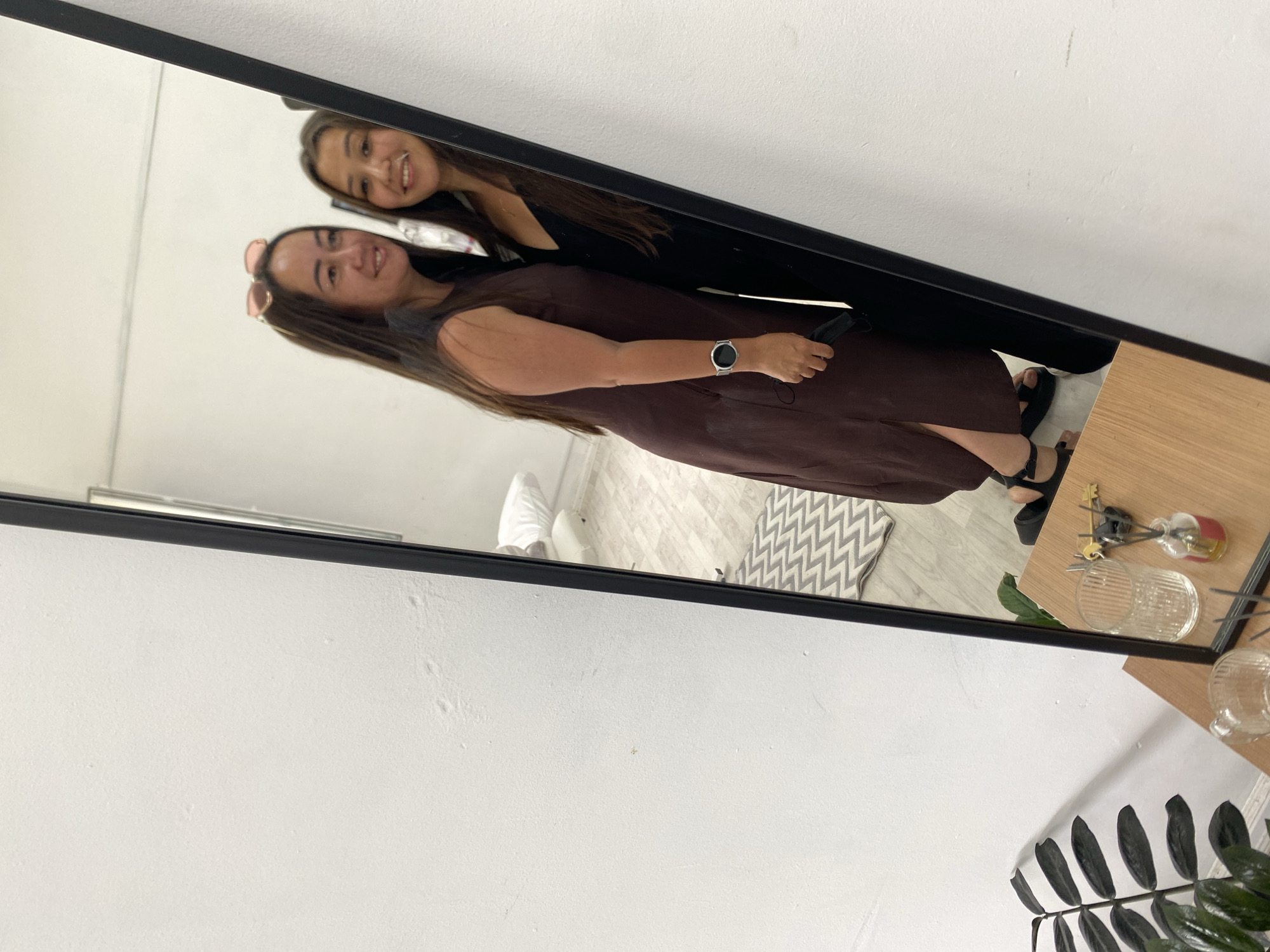 The image shows a reflection in a mirror of two women. The woman on the left is wearing a dark brown sleeveless dress and sunglasses pushed up on her head. She is wearing a smartwatch on her left wrist and holding a black face mask in her left hand. The woman on the right is wearing a black sleeveless dress and has long dark hair. They appear to be standing close together and looking at the camera. In the reflection of the room, a grey and white chevron patterned rug is visible on a wooden floor. In the foreground, a wooden table holds a clear glass, a small bottle with reeds, and keys. A plant with dark green leaves is positioned to the left of the table. The wall in the background appears to be white. There is no indication of the location being in Akyar, Russia.